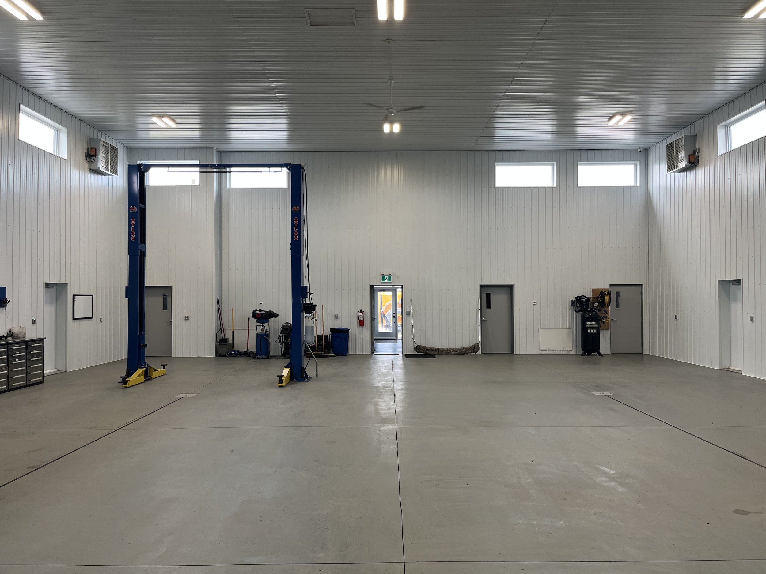 Empty industrial workshop with a blue vehicle lift, tools, and equipment along the walls, bright windows, and a ceiling fan.