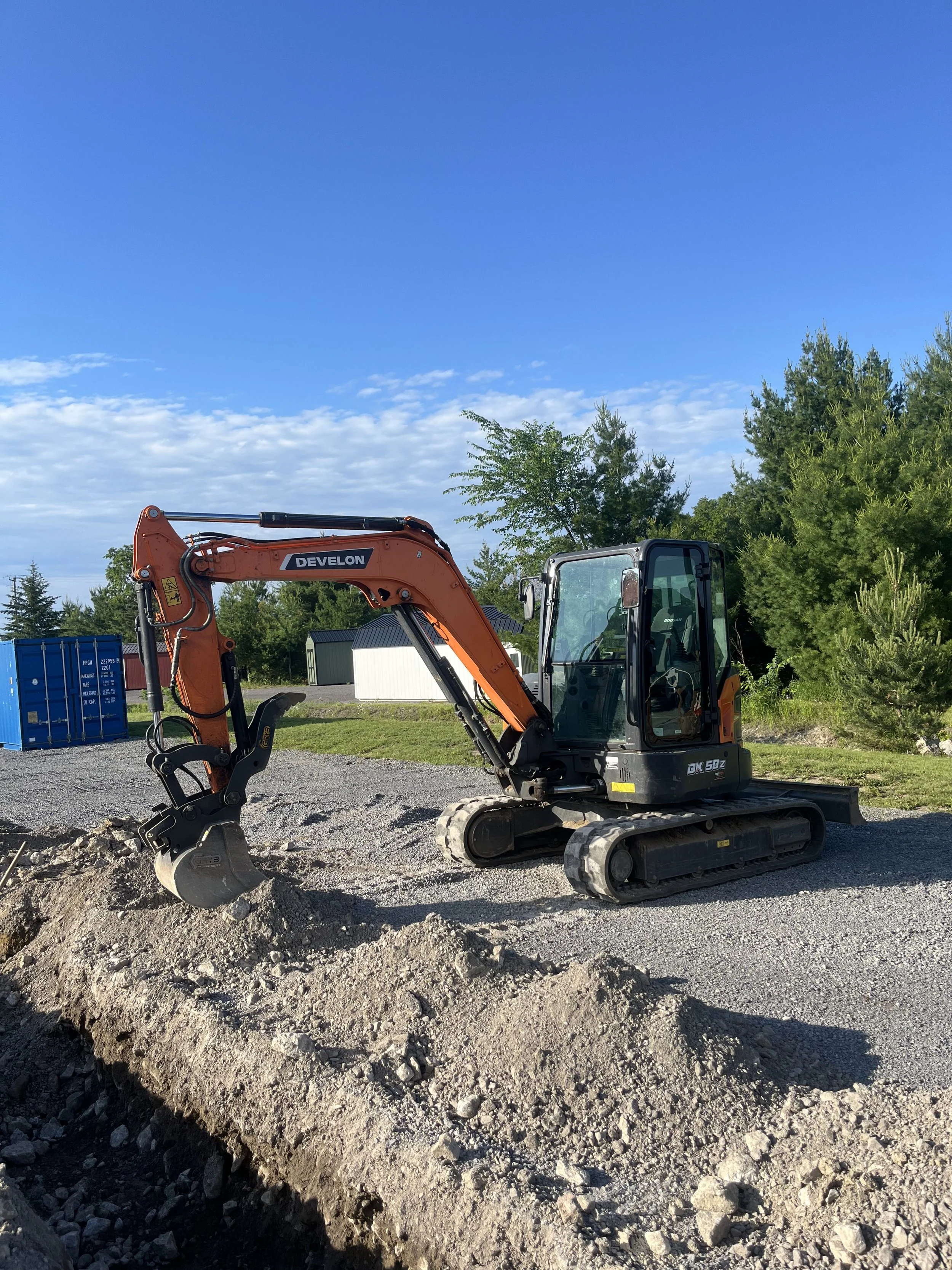 A small orange and black excavator with a bucket attached, working on a dirt surface at a construction site with trees and storage buildings in the background.