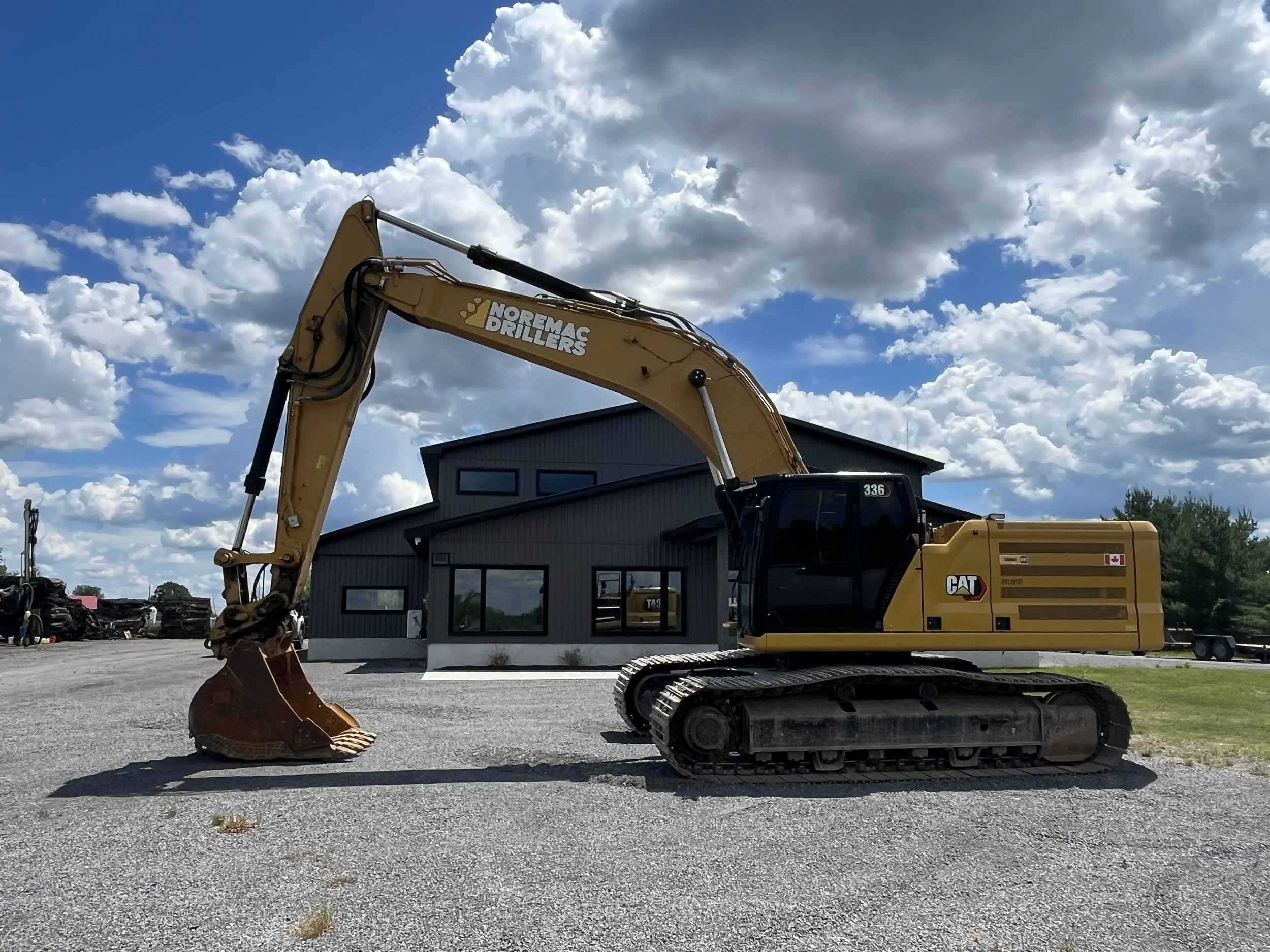 A yellow excavator with the brand name Noremac Drillers and Caterpillar, sitting on gravel in front of a dark gray building, under a partly cloudy sky.