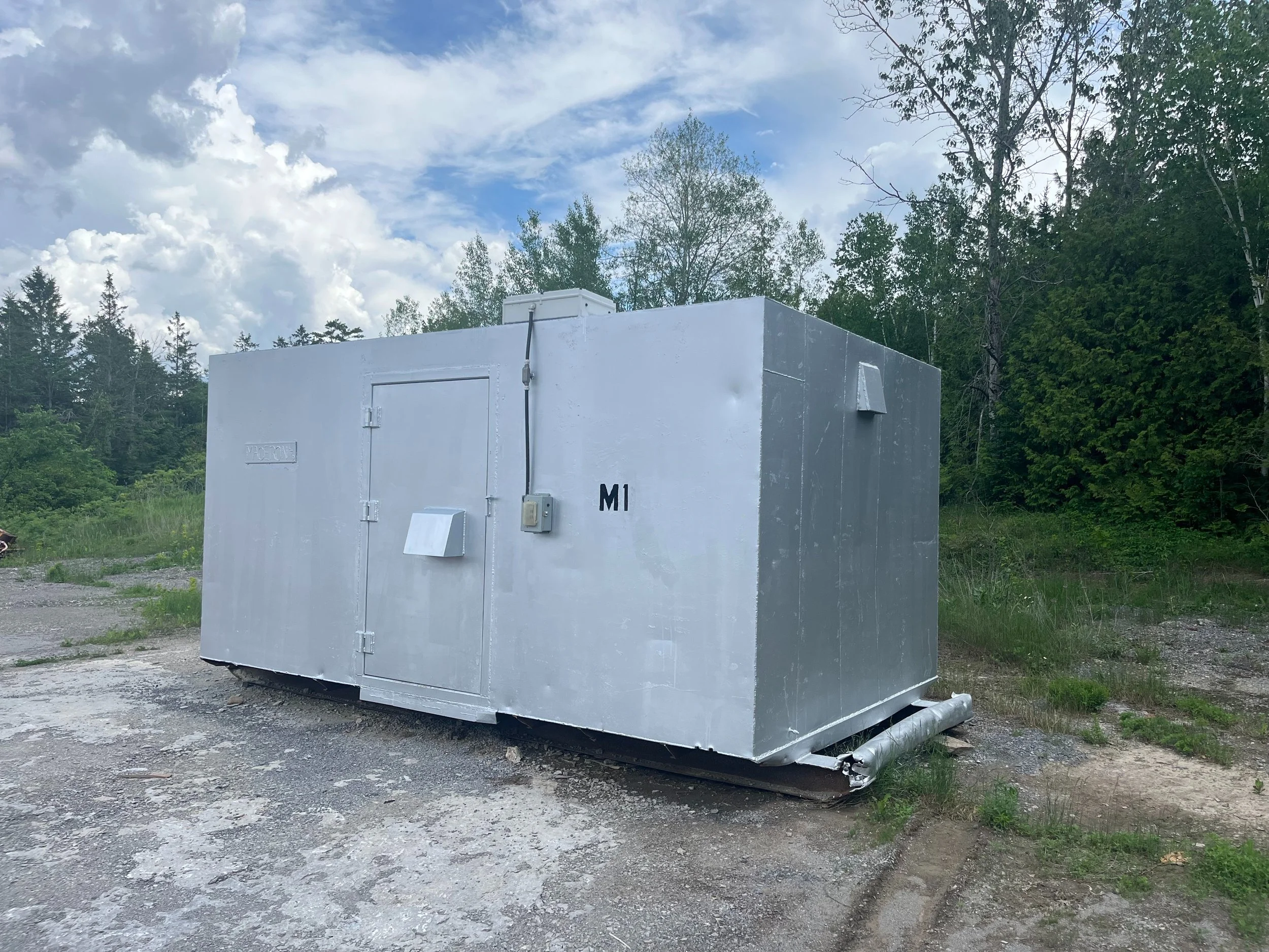 A white, rectangular container or equipment box with electrical connections, labeled "M1," placed outdoors on a gravel patch, with trees and a partly cloudy sky in the background.