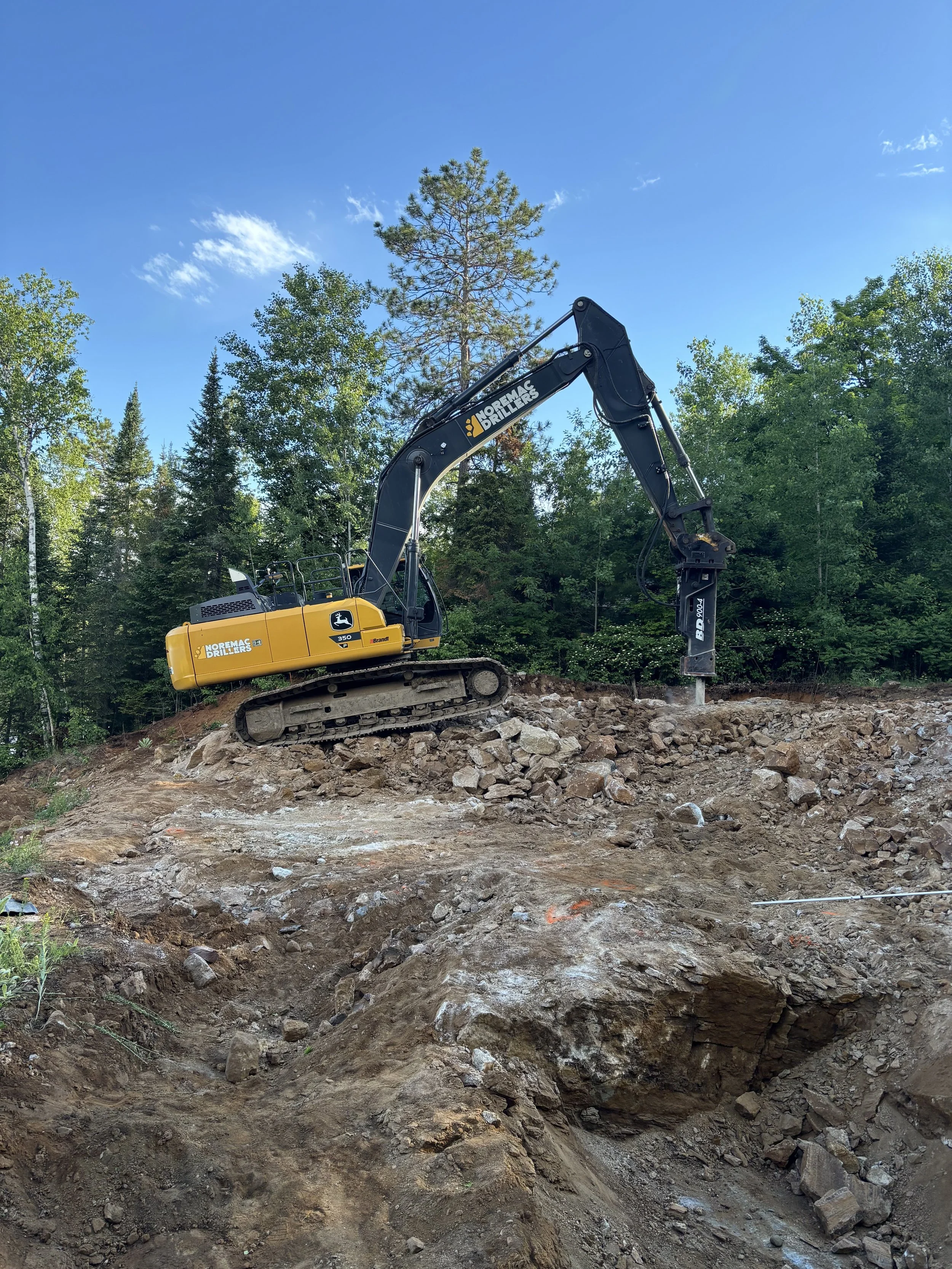 A construction excavator working on a rocky hillside in a forested area with green trees and a blue sky with some clouds.
