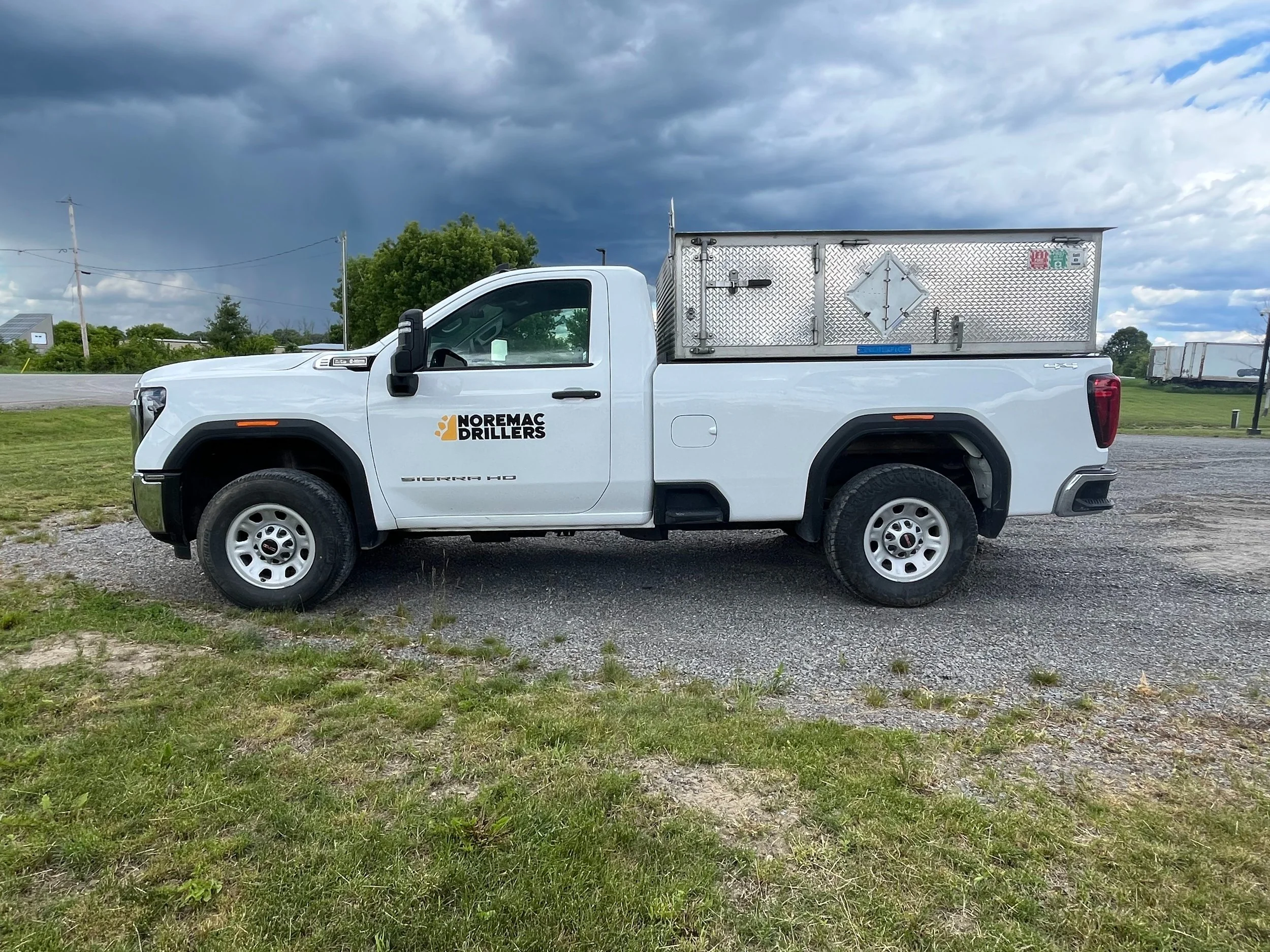 A white pickup truck with a metallic storage container on the bed parked on grass under a cloudy sky.