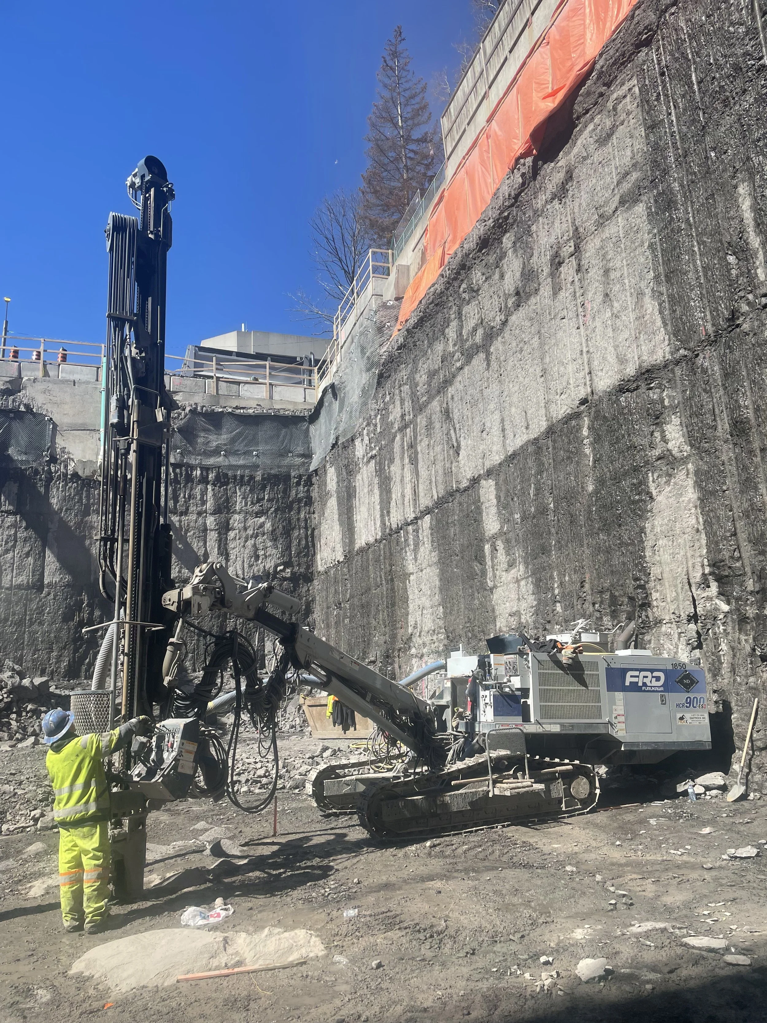 Construction workers operate heavy machinery at a construction site on a rocky cliff, with safety gear and a clear blue sky in the background.