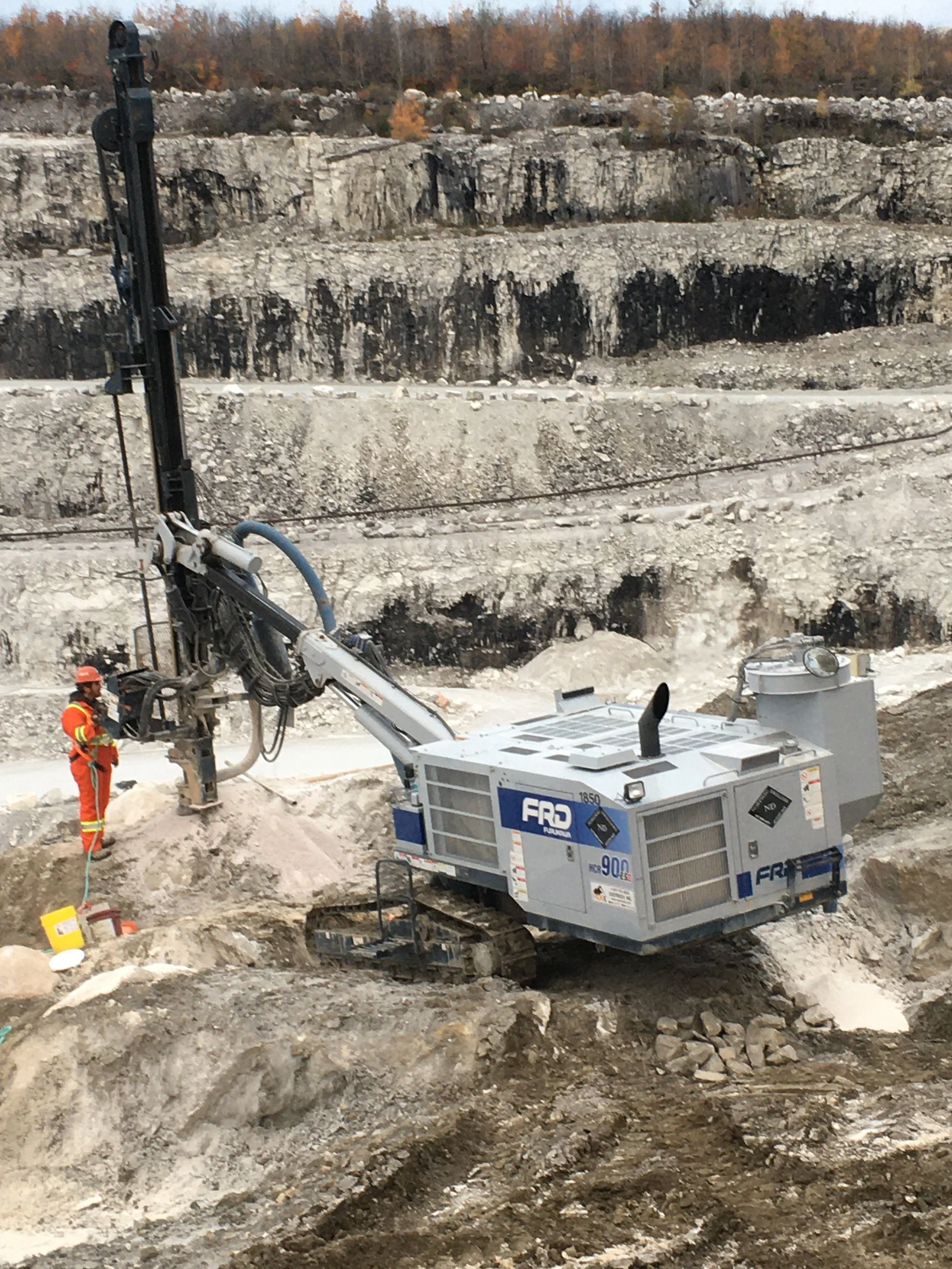 Construction site with a large drilling machine and two workers wearing orange safety gear. Excavated earth and rocks are visible in the foreground, with a rocky hillside and trees in the background.