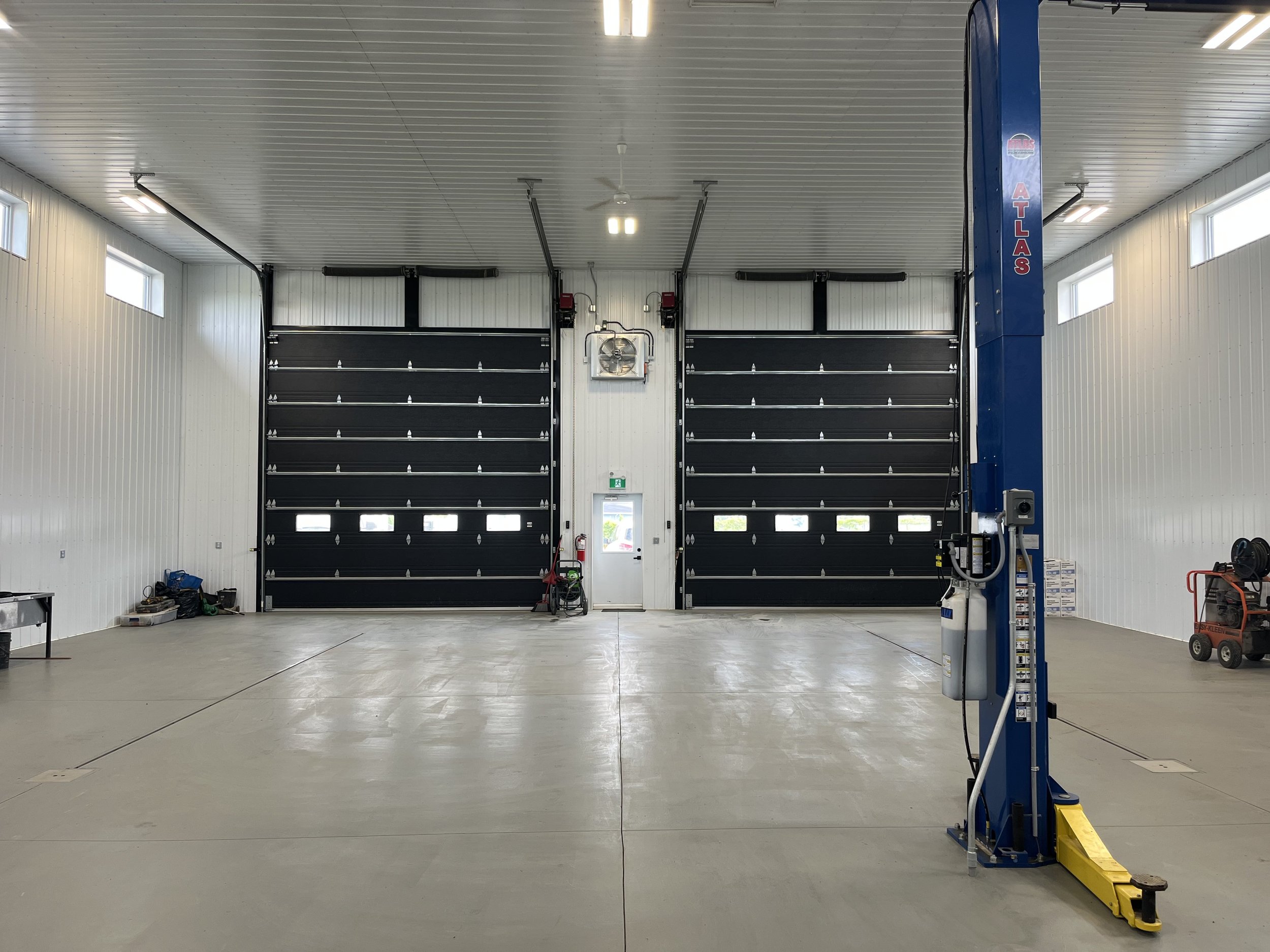 Inside an empty garage or industrial building with high white paneled walls and ceiling, fitted with small rectangular windows for natural light. There are two large black sectional garage doors at the back, with an emergency exit door beneath the center. Equipment, tools, and supplies are scattered on the floor and shelves on the sides. A blue vehicle lift or hoist stands on the right side of the image.