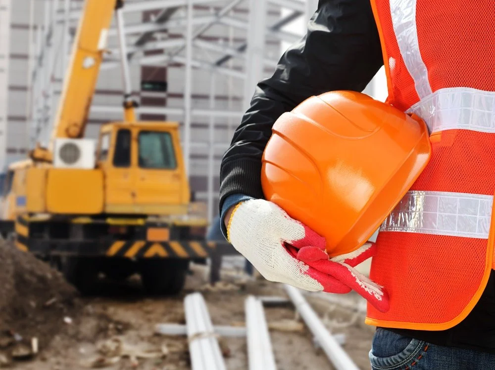 Construction worker holding an orange safety helmet at a construction site with a yellow crane and building framework in the background.