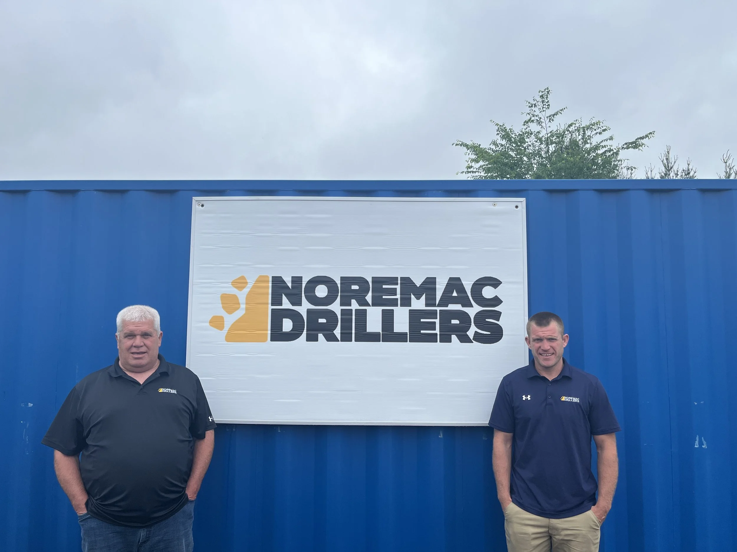 Two men standing in front of a blue wall with a sign that reads "NOREMAC DRILLERS" and logo. The man on the left wears a black polo shirt with the logo, and the man on the right wears a navy blue polo shirt with the logo.