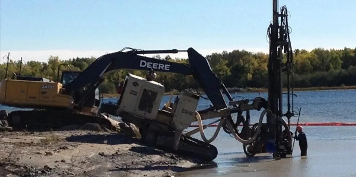 An excavator operating in water near a shoreline, with a worker standing nearby, and trees in the background.