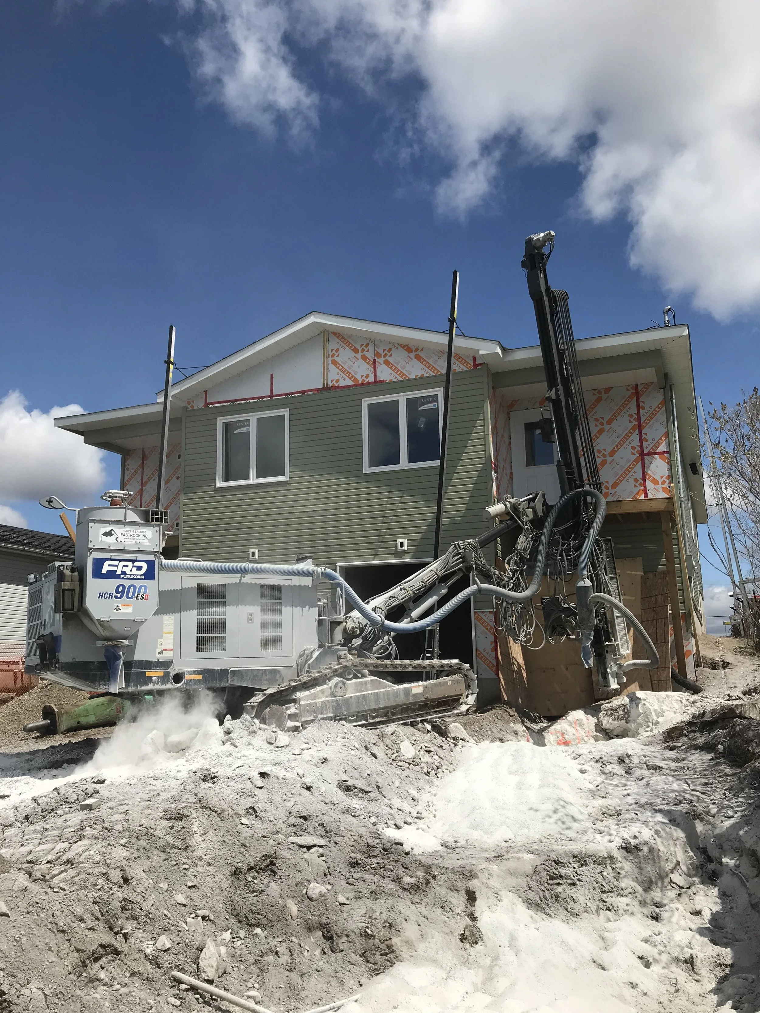 Construction site with a house being built and a large excavator on muddy ground, blue sky with clouds.