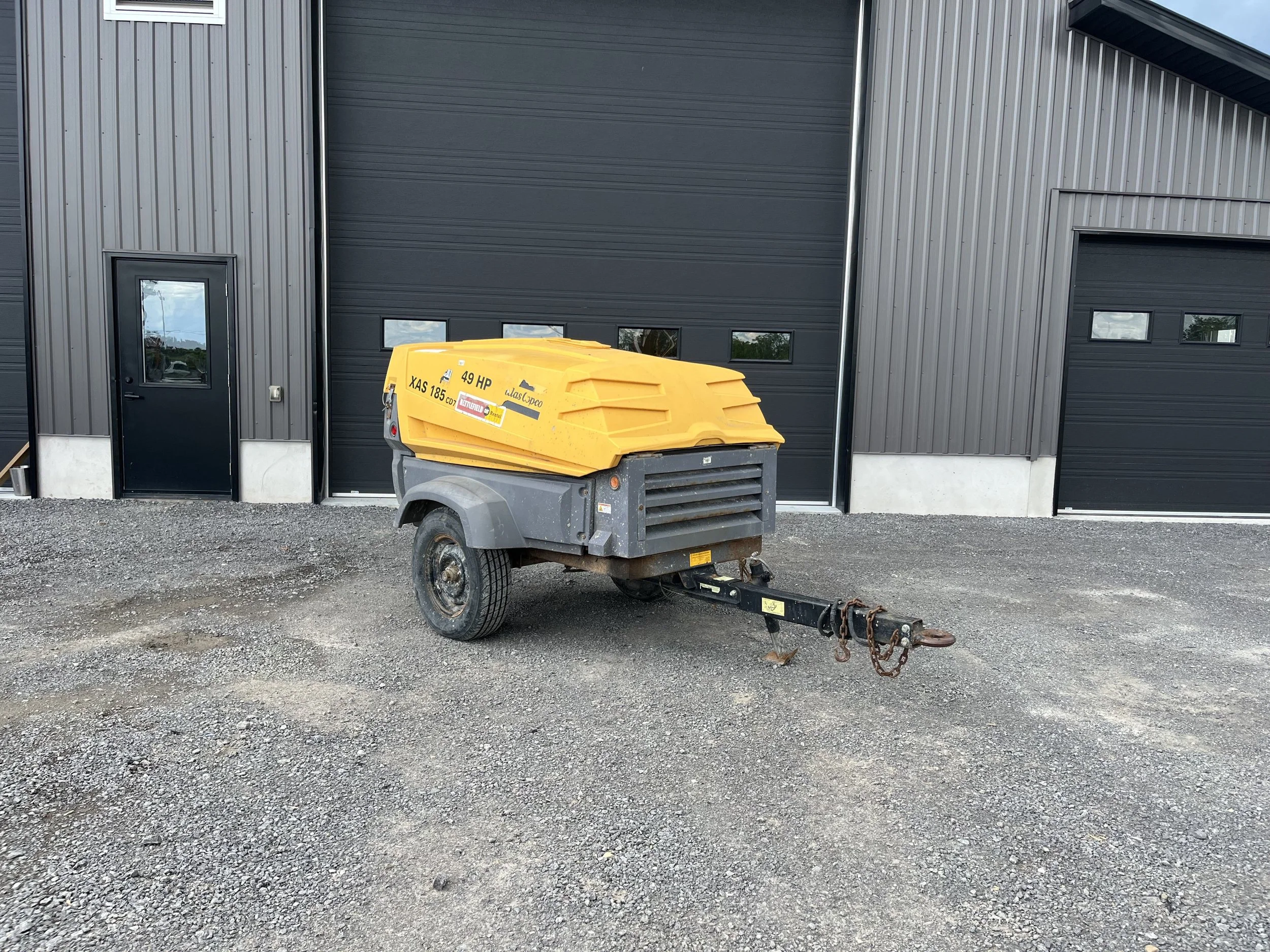 A yellow portable air compressor on a trailer hitch in front of a modern building with black and gray metal siding and small windows.