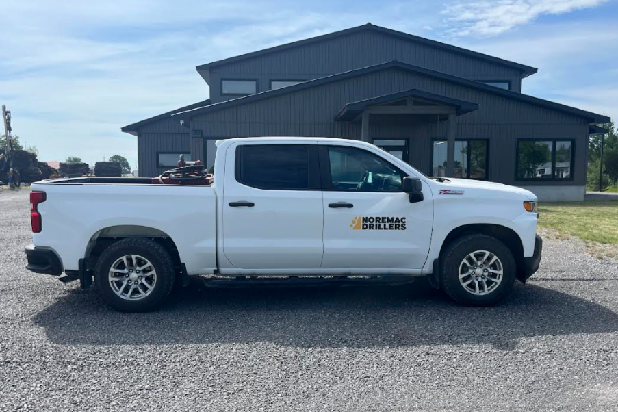 White pickup truck with 'Noremac Drillers' logo parked on gravel in front of a modern gray building with large windows and a blue sky.