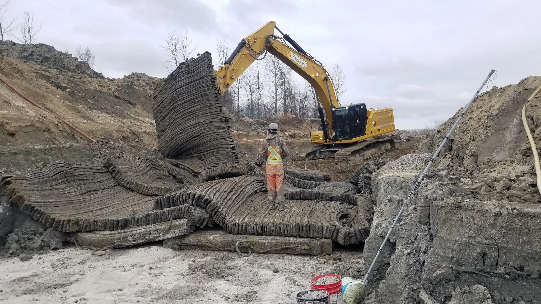 A construction site with a large excavator lifting a layered, petrified log or fossil. A worker in safety gear stands nearby, observing the excavation. The site has dirt mounds and construction equipment scattered around.