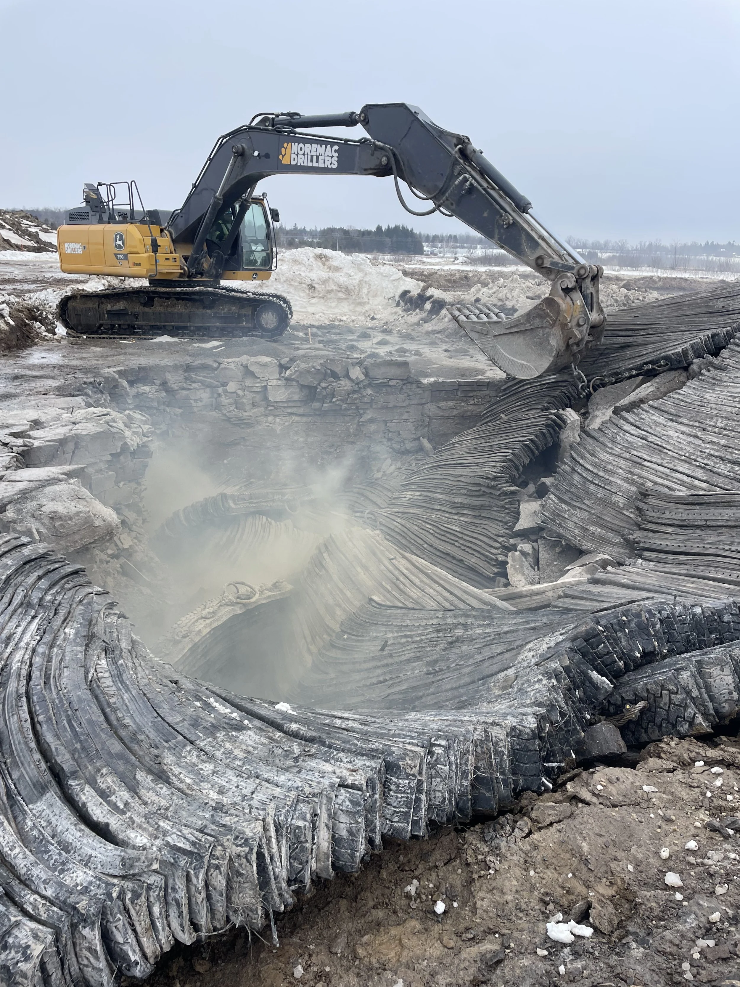 Excavator digging in the ground with snow and rocky terrain in the background.