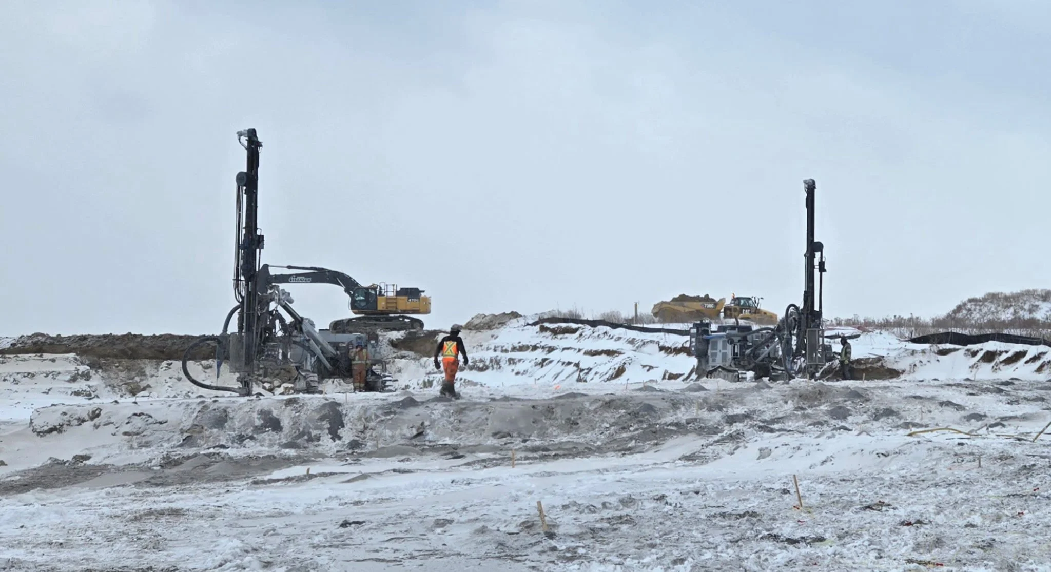 Construction workers operating heavy machinery on a snowy site, with snow-covered ground and overcast sky.