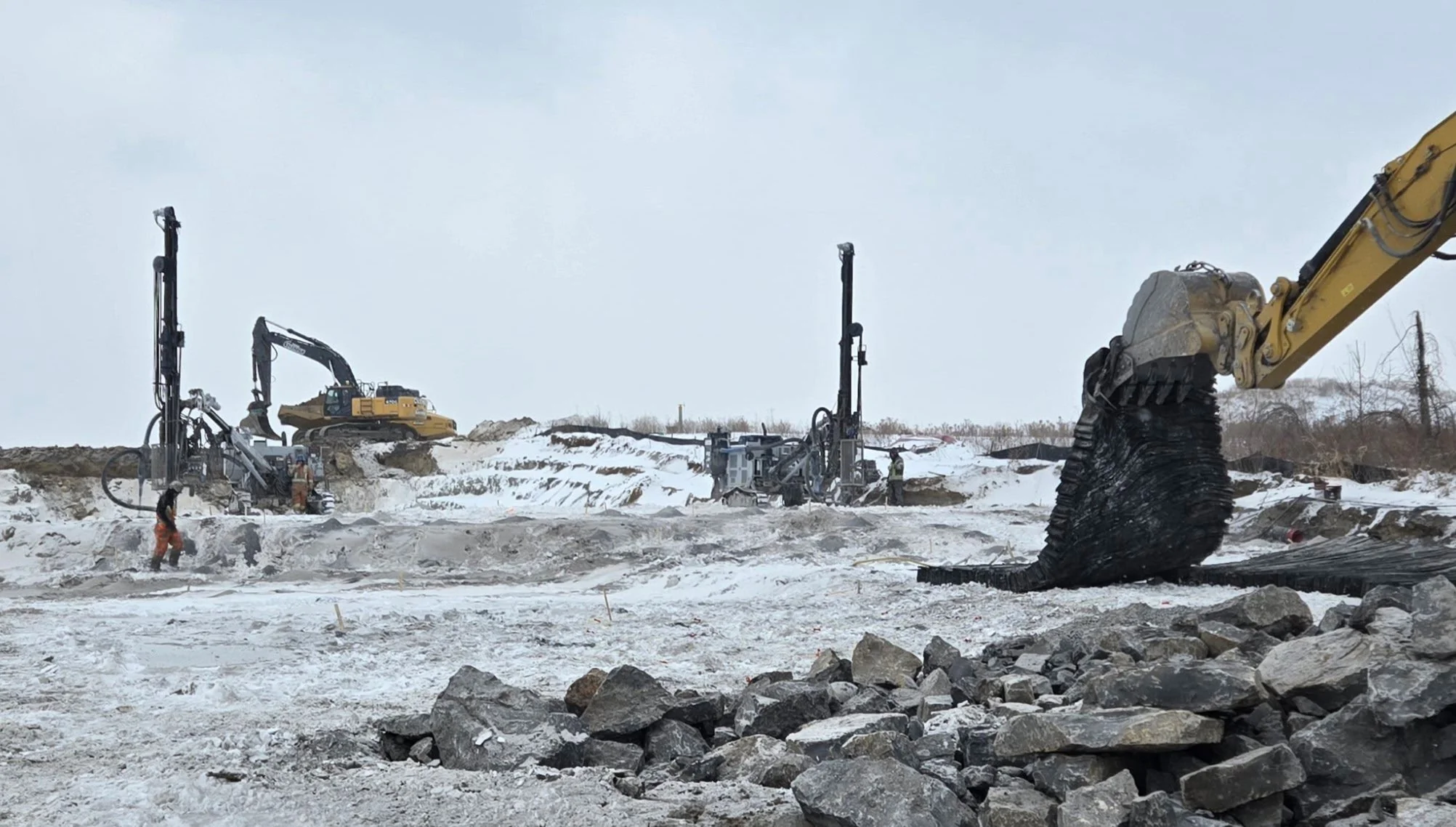 Construction site with large excavator and drilling equipment on snow-covered ground, workers nearby, in a cold, possibly winter weather setting.