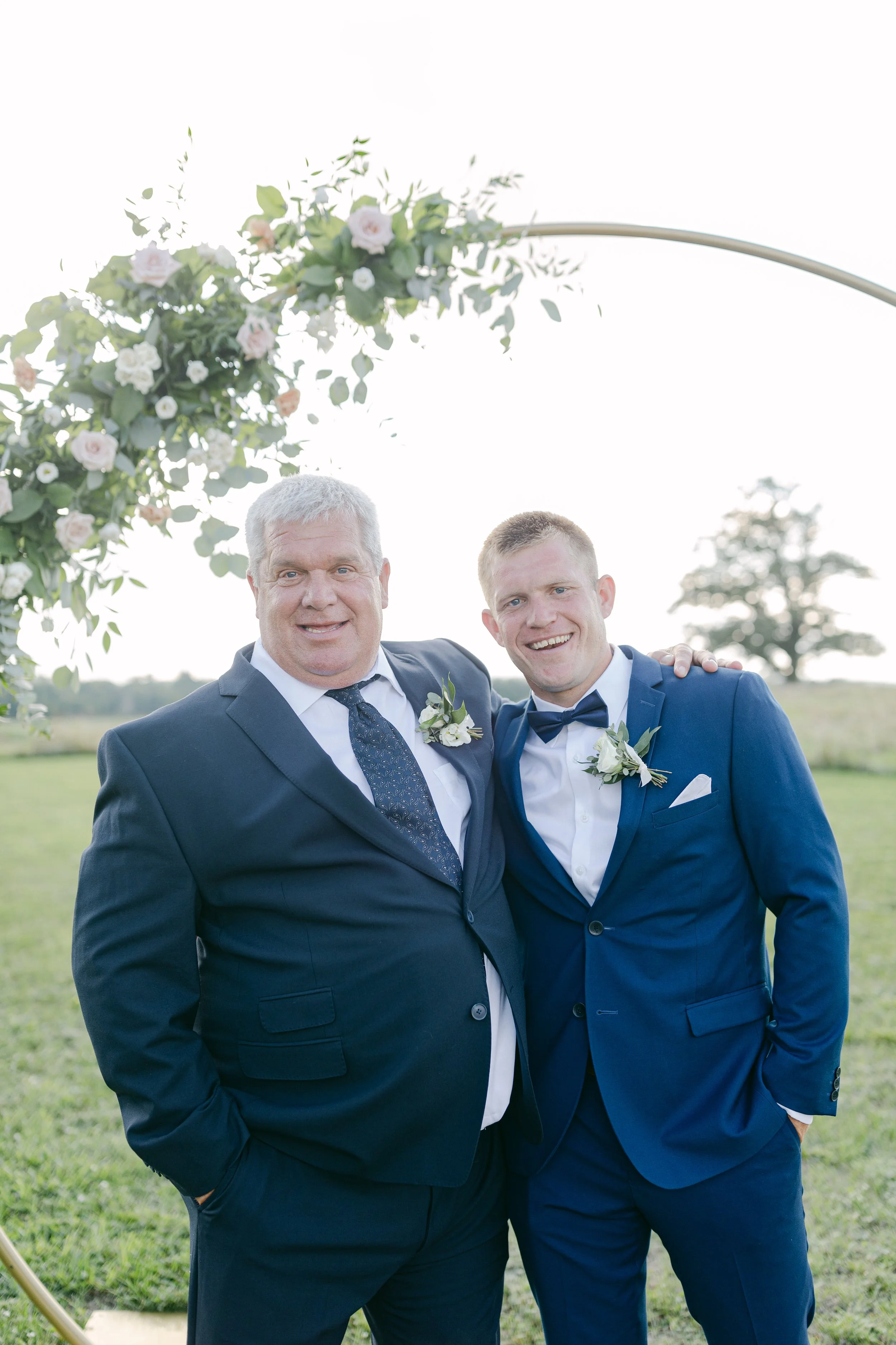 Two men in suits smiling at an outdoor wedding ceremony, standing under a floral arch with greenery and pale pink and white roses, in a grassy field during daytime.
