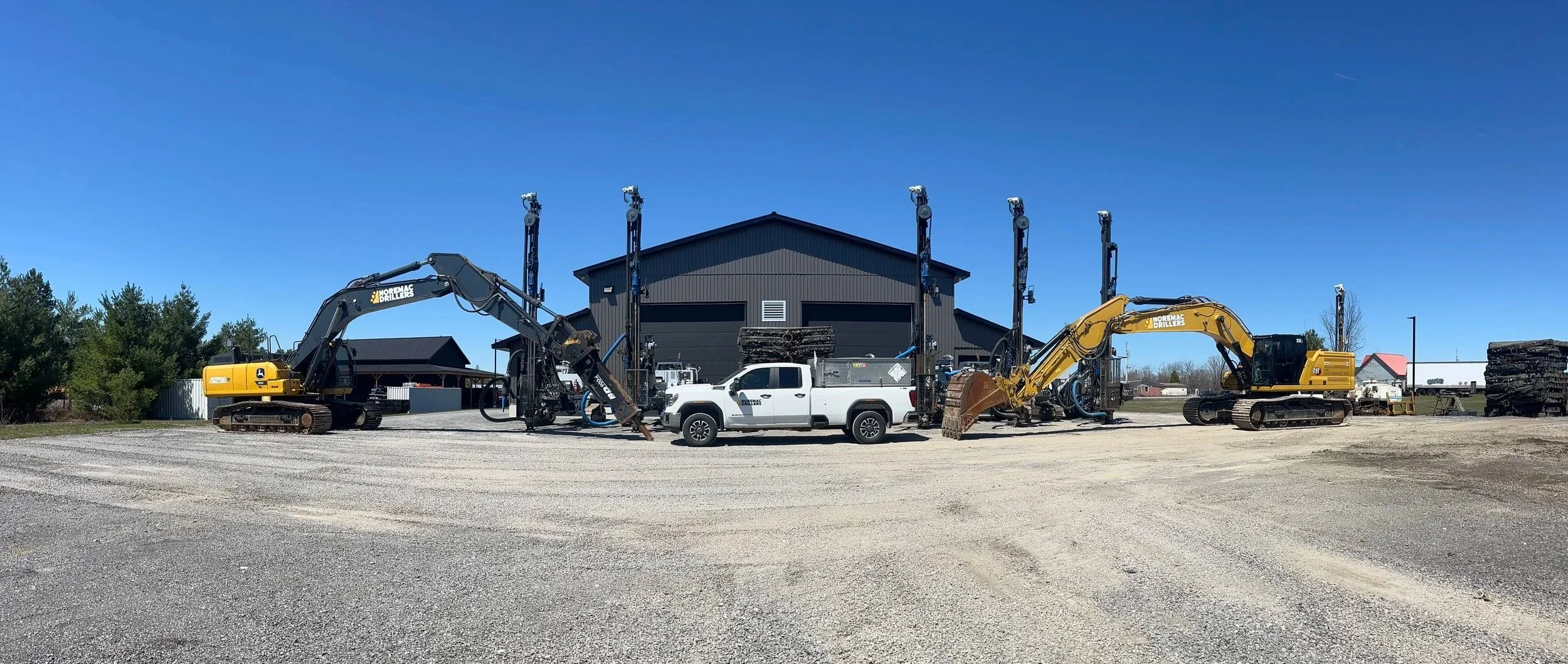 Construction equipment including two excavators and a pickup truck parked on gravel in front of a black industrial building, under a clear blue sky.