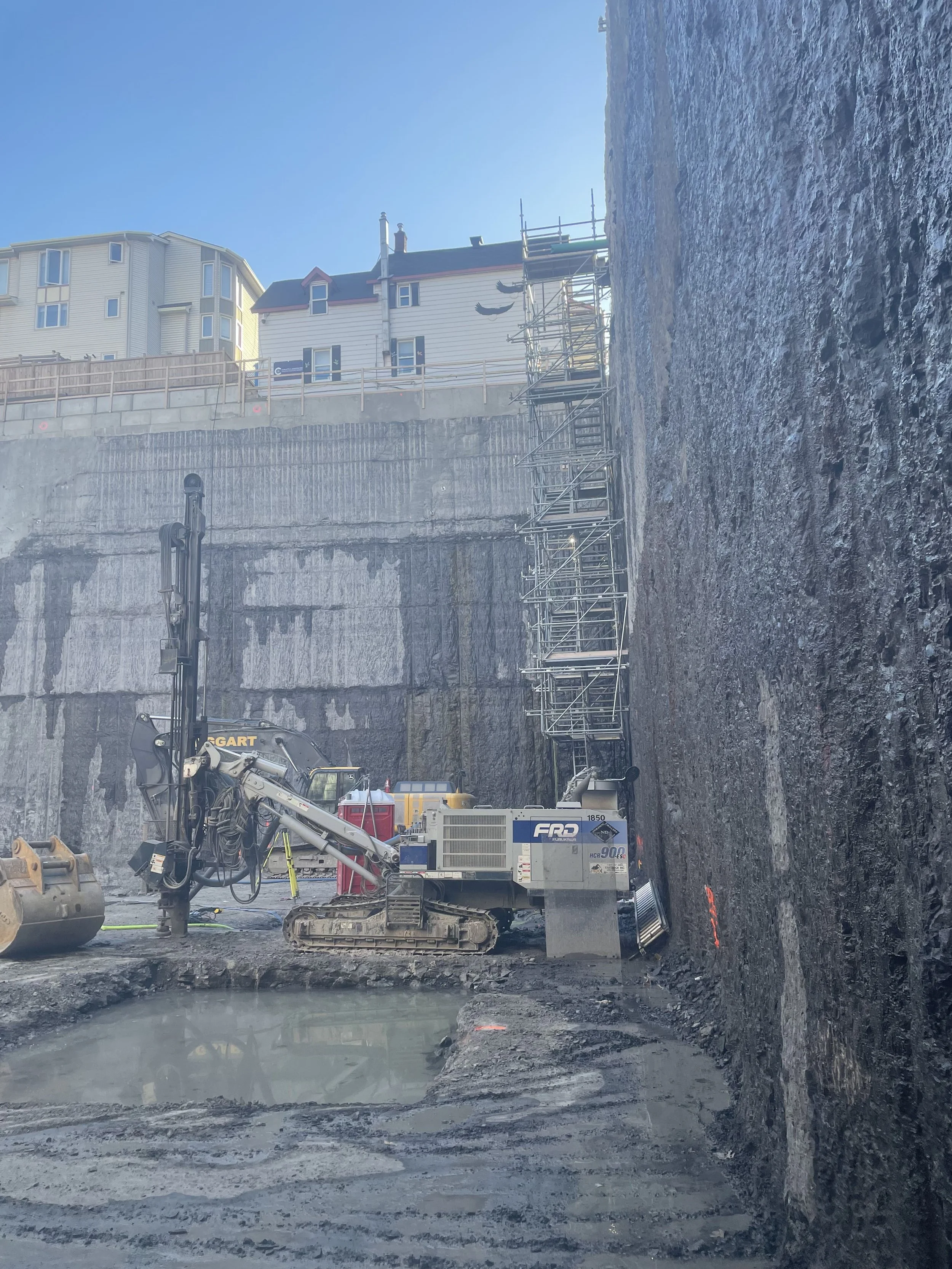 Construction site with machinery, scaffolding, and partially excavated area, with buildings and blue sky in the background.