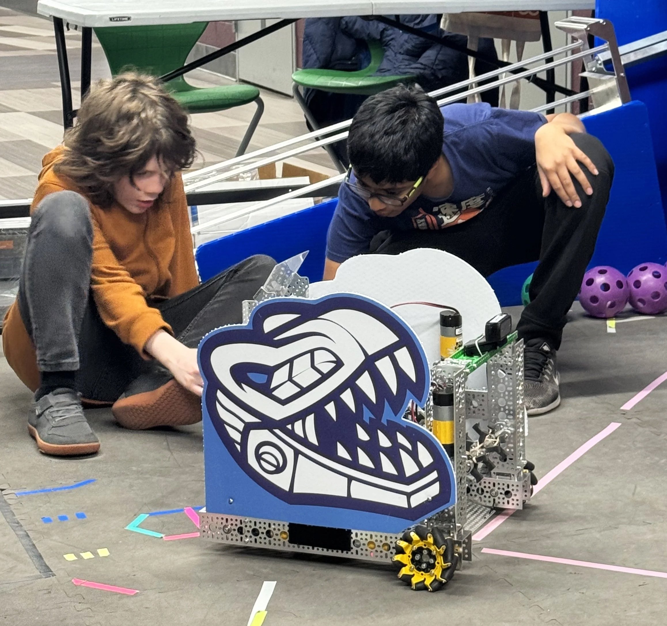 Two people working with a robot or mechanical device on a table at an industrial or technical event. There are colorful tape markings on the floor and a set of purple and yellow wheels or gears on the side of the device.