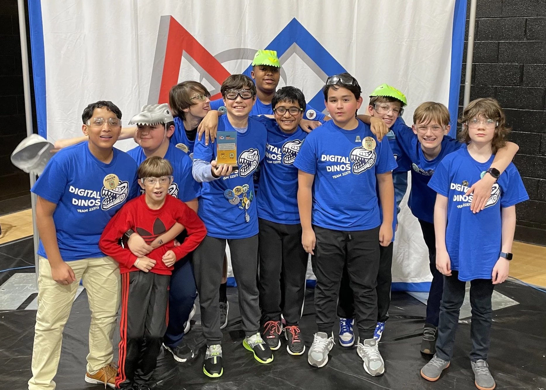 A group of ten children, wearing blue 'Digital Dinos' team shirts, are posing together at a robotics or STEM event. They are smiling, with some wearing safety goggles or dinosaur hats. The background features a white curtain with a colorful geometric logo. The children appear to be celebrating a victory or achievement.