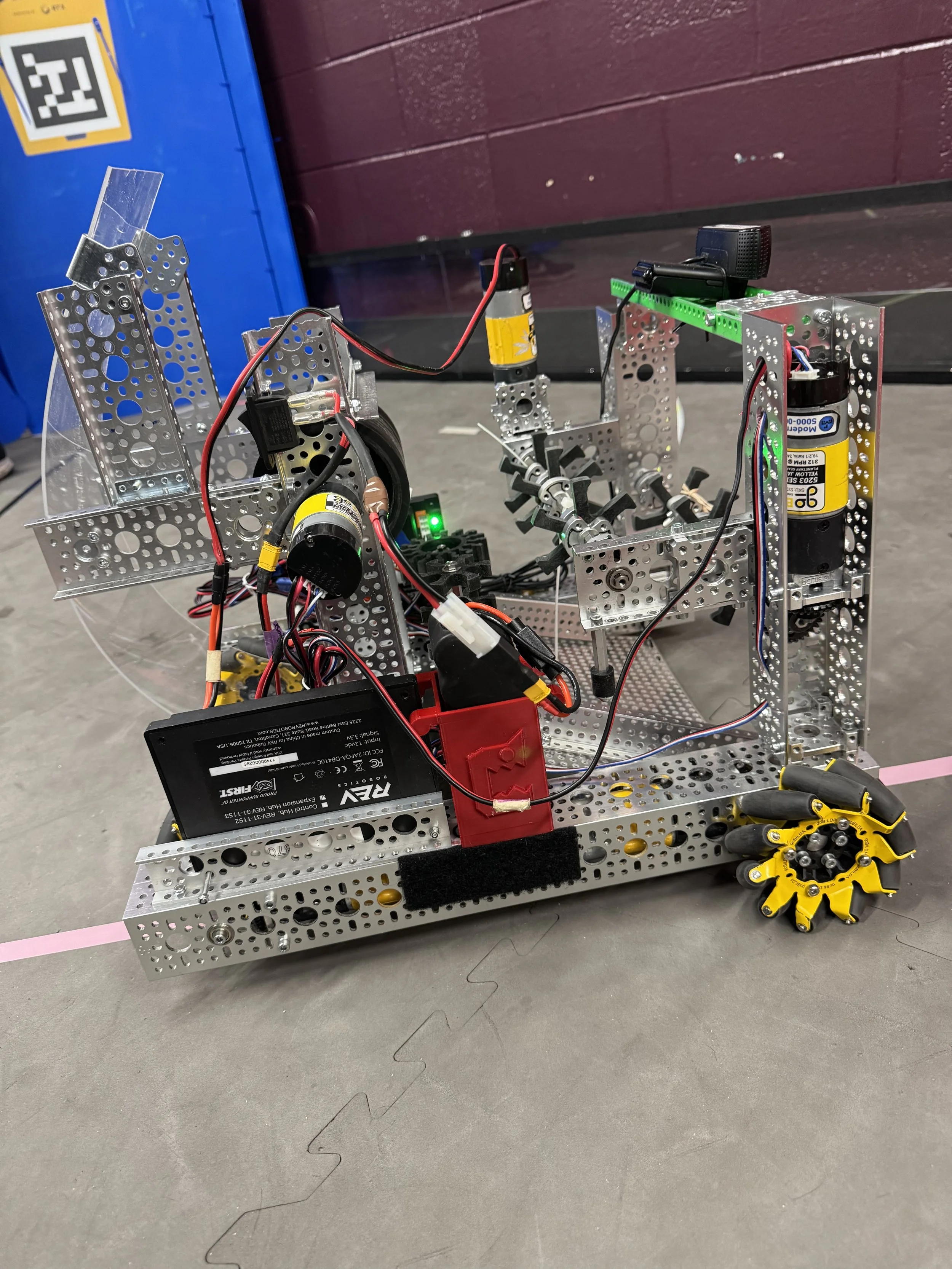 A small robot built with metal parts, wires, and electronic components on a gray floor, with a blue and maroon background.