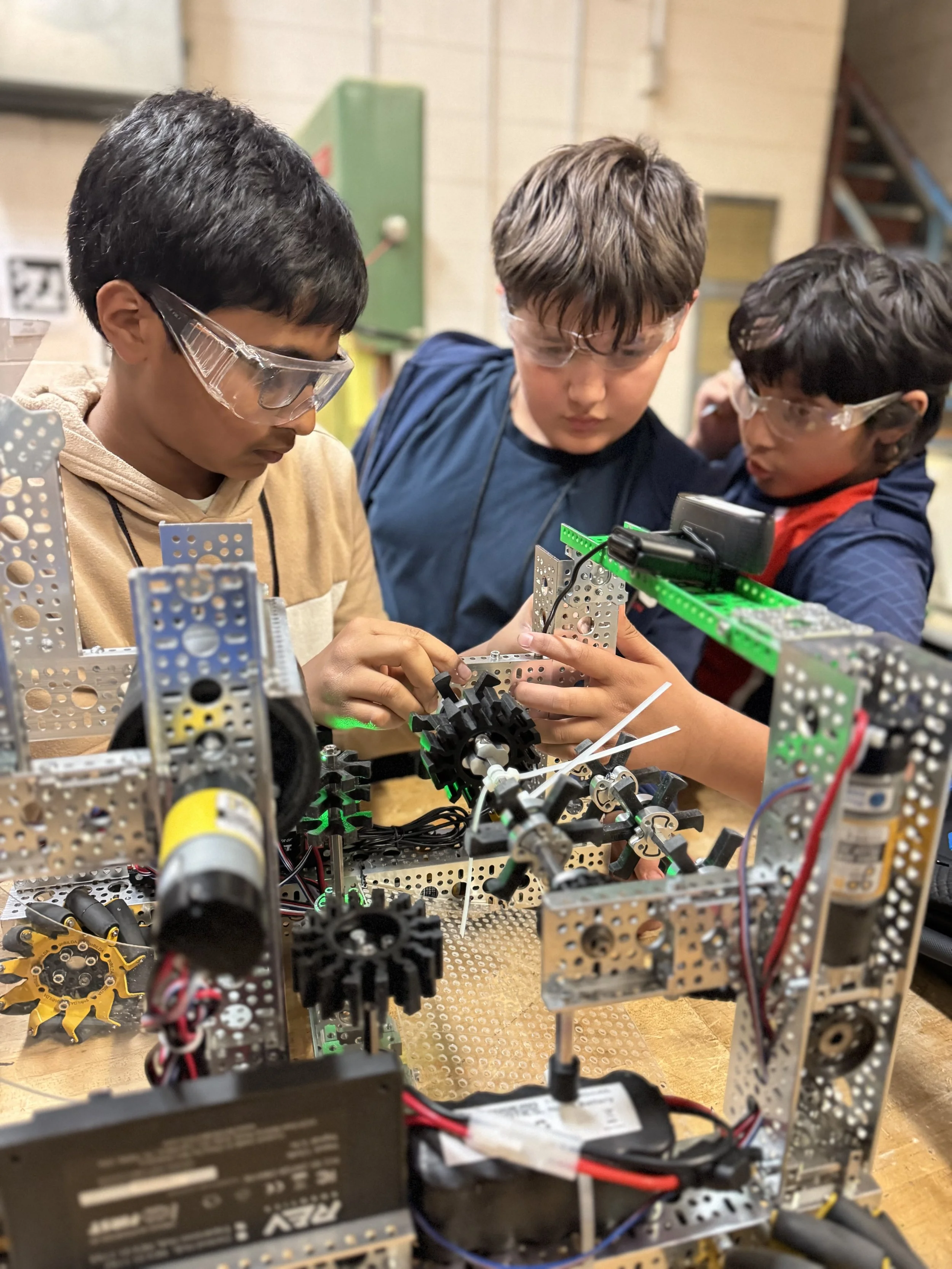 Three boys wearing safety glasses working on a robotics project with metal parts, wires, and gears in a workshop or classroom setting.