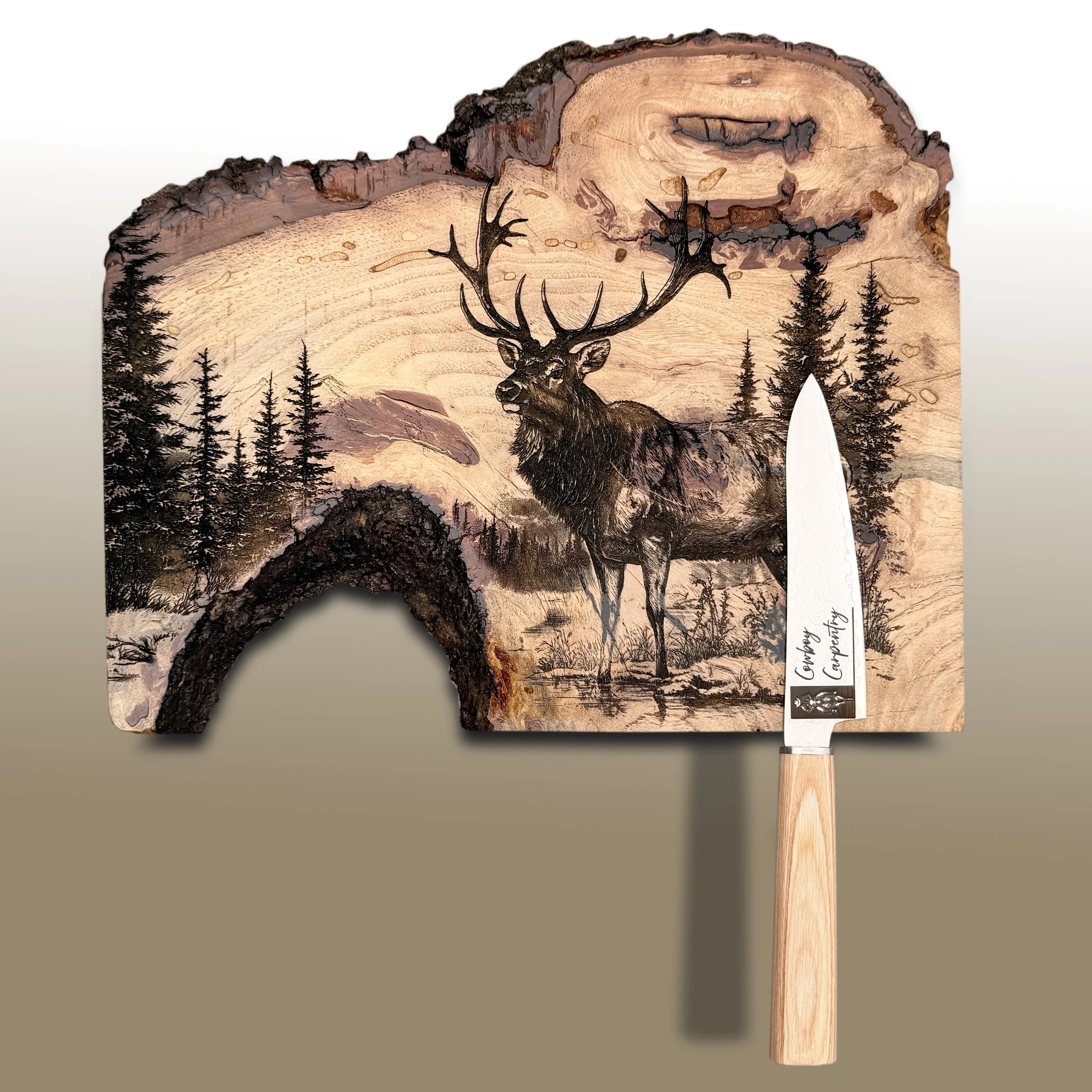 A wooden cutting board with mountain landscape and forest scene, featuring a carved stag with large antlers and a forest of tall pine trees, mounted on a handle with a white tag.