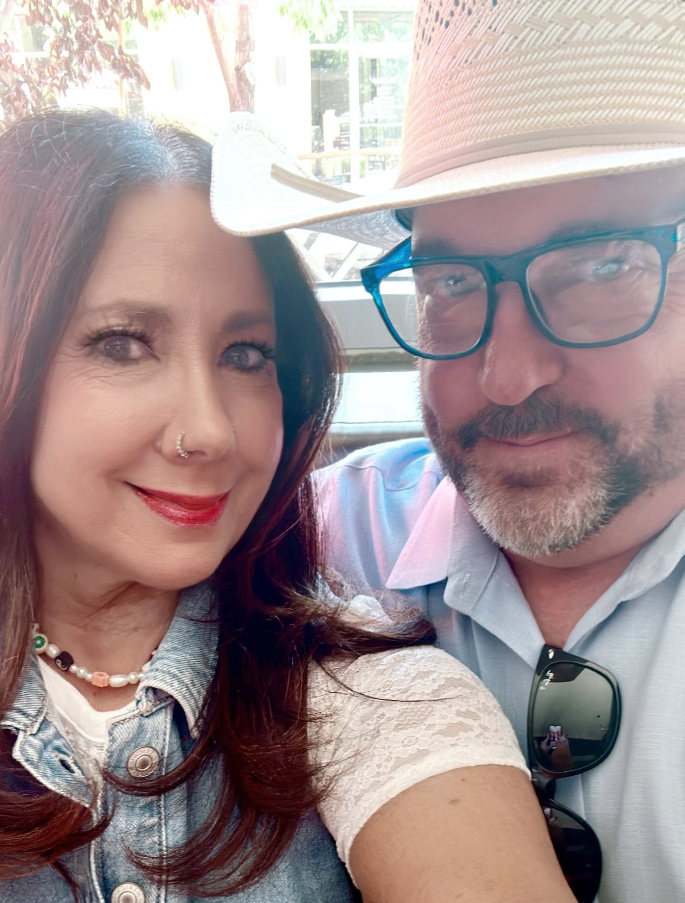 A man and a woman taking a selfie together. The woman has long reddish-brown hair, makeup, and is wearing a necklace, a denim vest, and a beige blouse with lace sleeves. The man has gray hair, a beard, glasses, and is wearing a wide-brimmed straw hat, a light-colored shirt, and sunglasses hanging from his shirt.