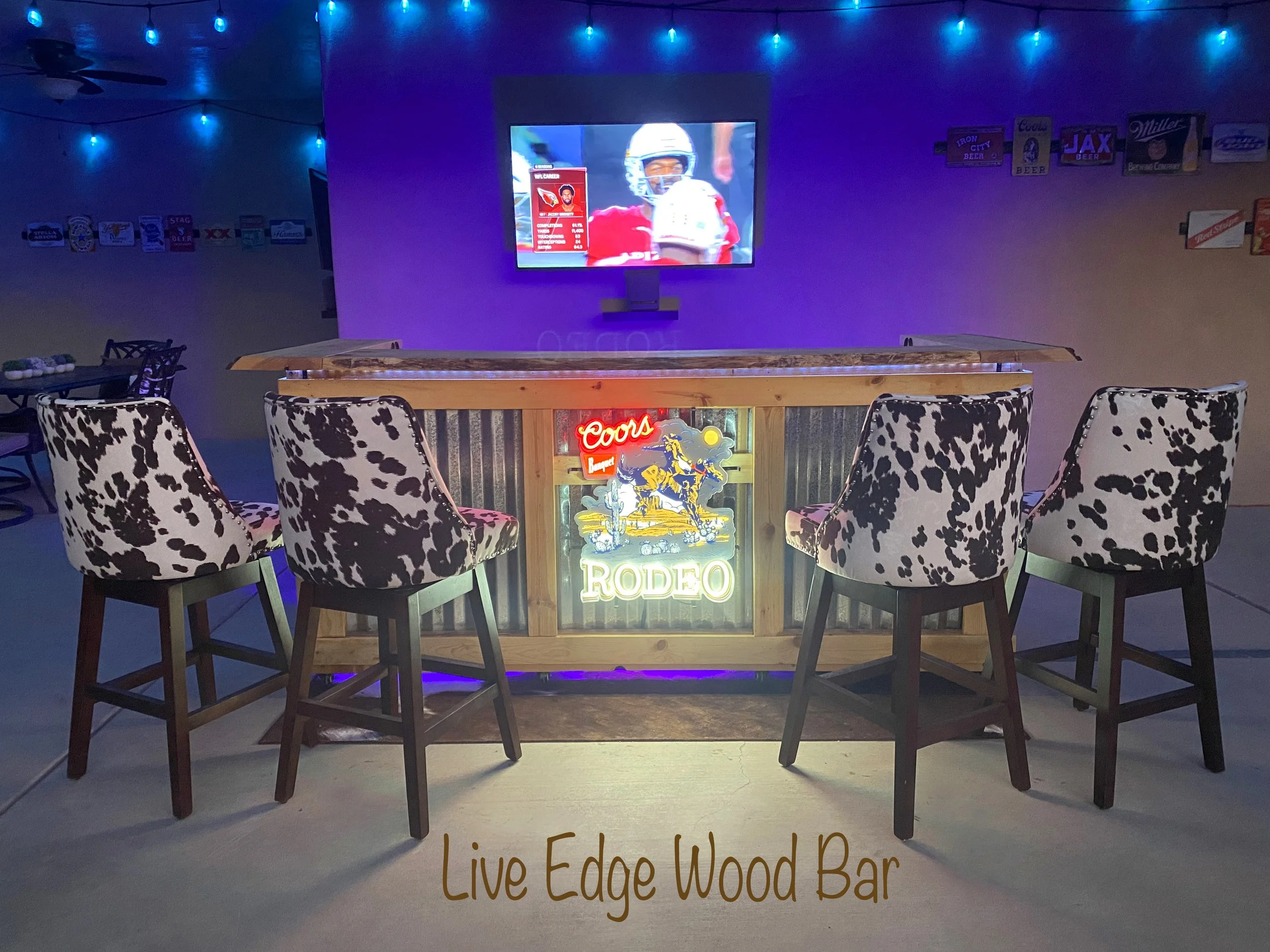 Bar with a television mounted on the wall displaying a football game, neon sign reading "Coors" and a sign below it that says "Live Edge Wood Bar" with cow print upholstered chairs around a wooden counter, blue string lights hanging from the ceiling,
