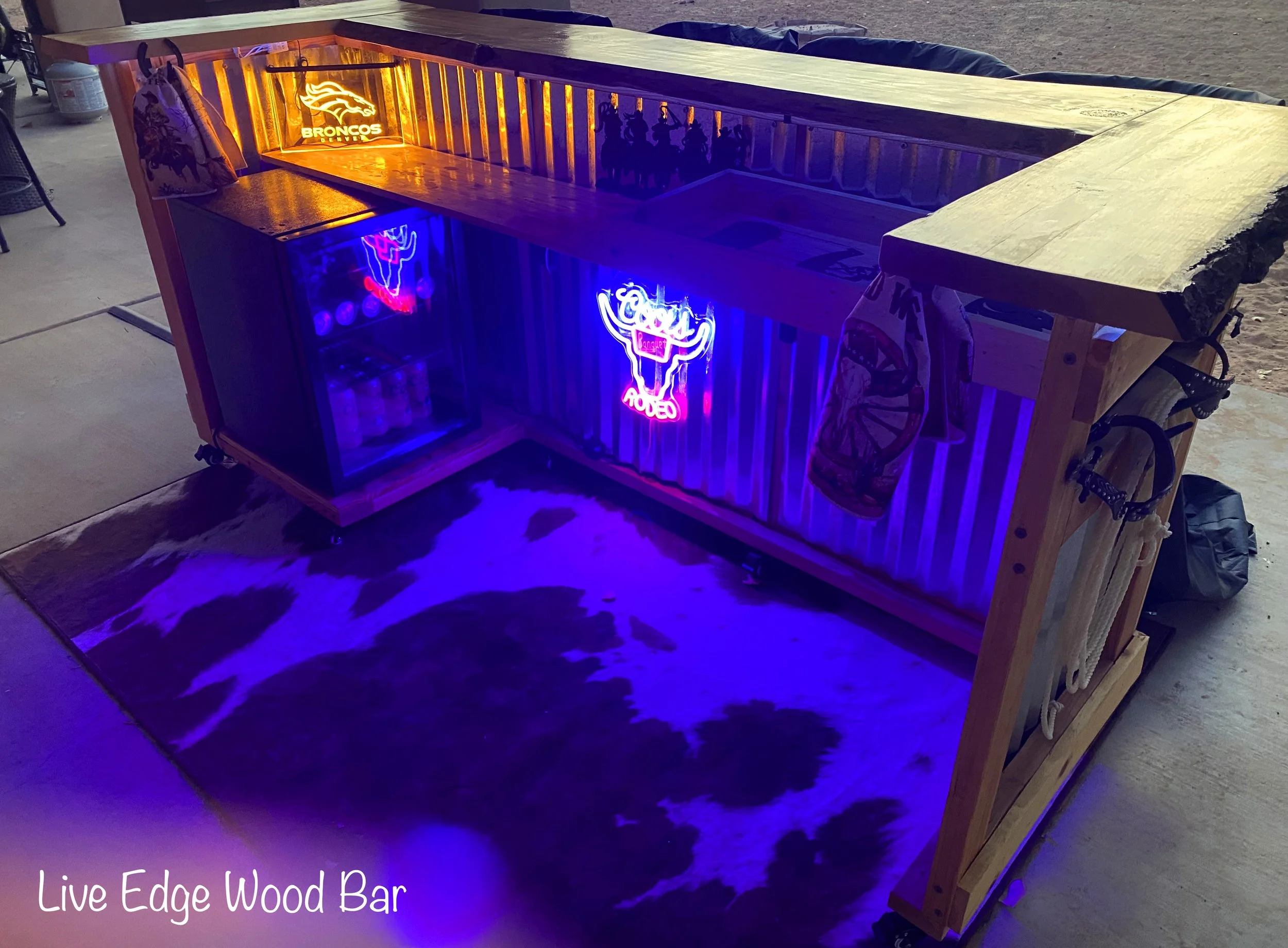 A wooden bar with neon signs and a mini fridge, illuminated with purple and yellow lighting, situated on a cowhide rug in a garage or workshop setting.