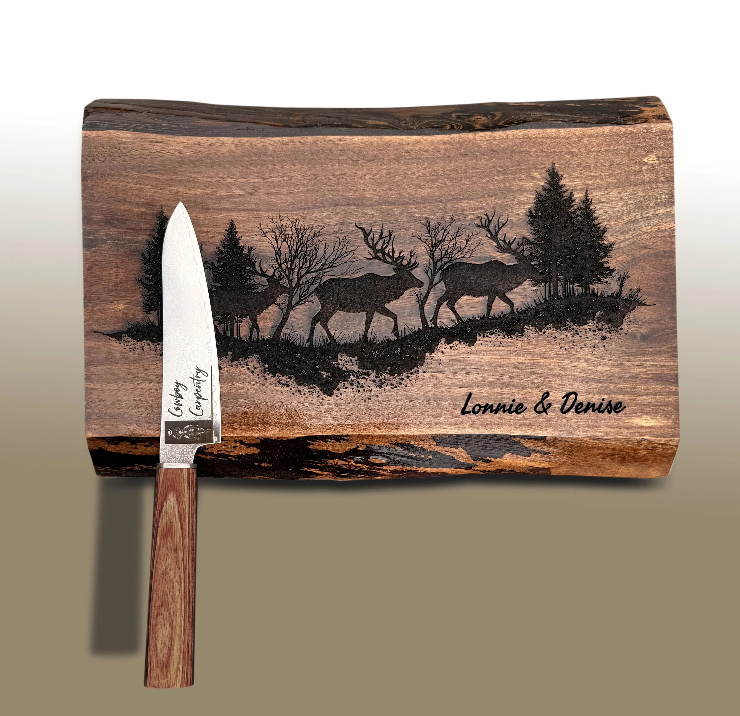 A wooden carving with a mountain scene featuring a group of moose, trees, and a sunset sky. Personalization is seen with the names 'Lonnie & Denise' and a white blade with black text that says 'Congratulations.'