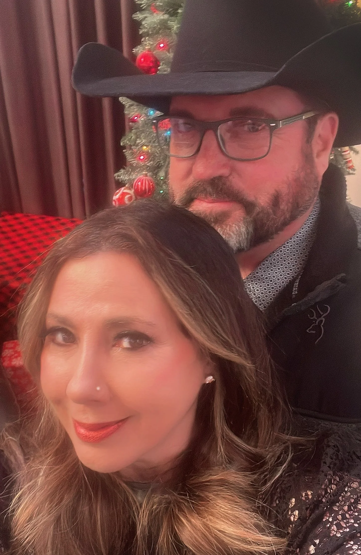 Close-up of a woman with brown wavy hair and makeup, and an older man wearing glasses, a black cowboy hat, and a patterned shirt, standing in front of a Christmas tree decorated with red ornaments and multicolored lights.