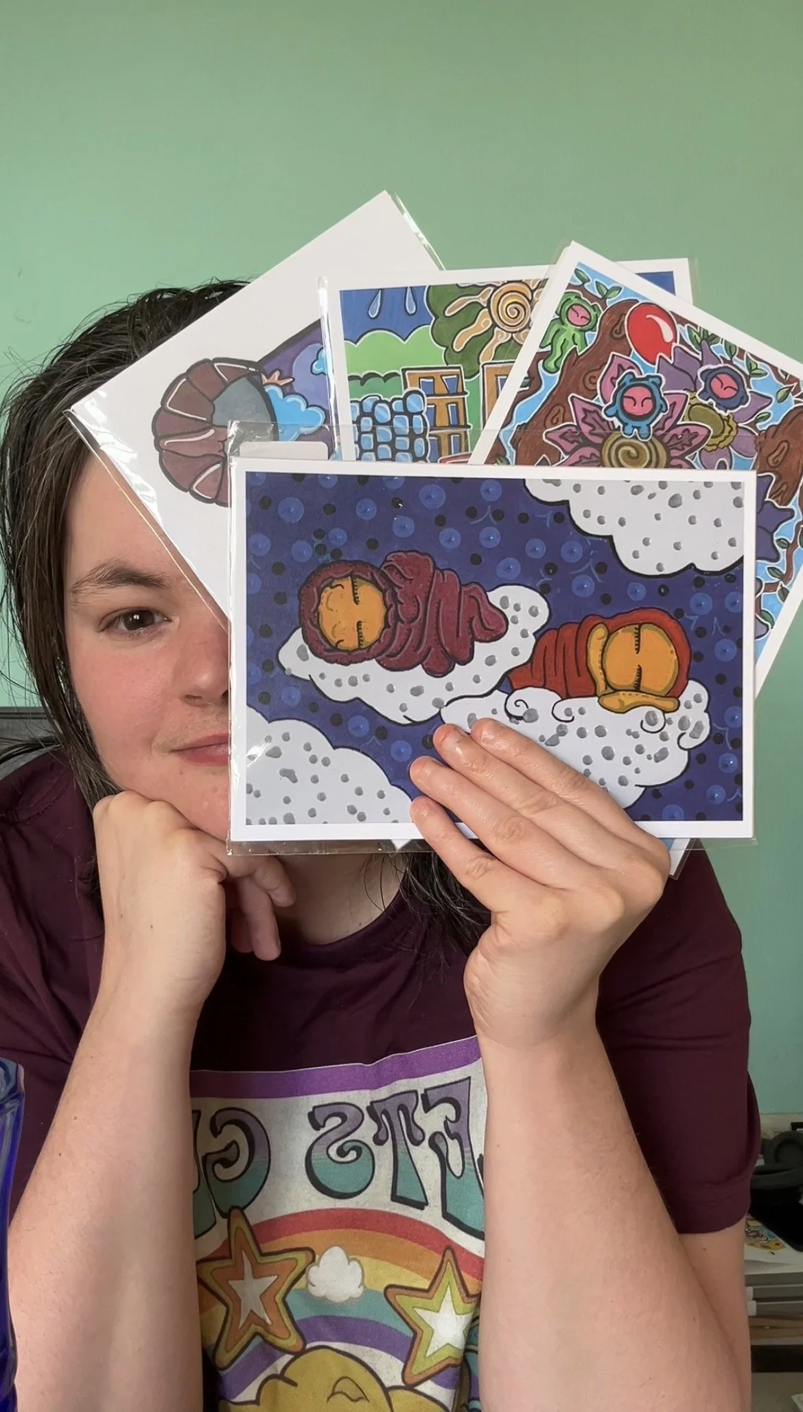 A woman holding three colorful cartoon artwork prints, partially obscuring her face against a light green wall.