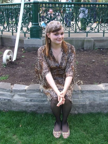 A young woman sitting on a stone border in a park, wearing a leopard print dress and matching headband, with black tights and floral shoes, smiling at the camera.