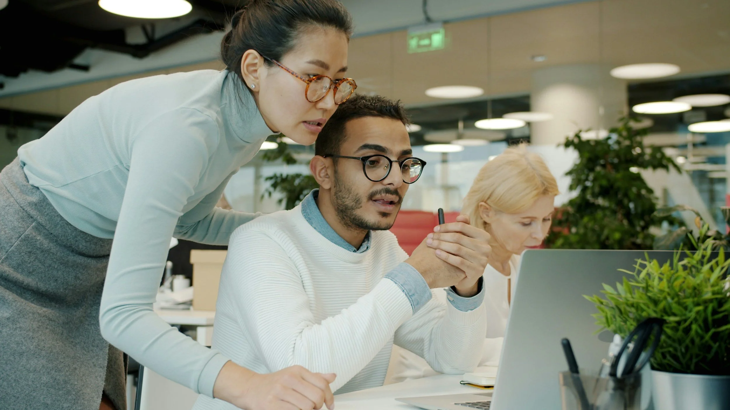 A woman wearing glasses and a white sweater leans over a seated man with glasses and a white shirt, both looking at a laptop screen in an office environment. A woman with blonde hair is sitting at the table in front of them.