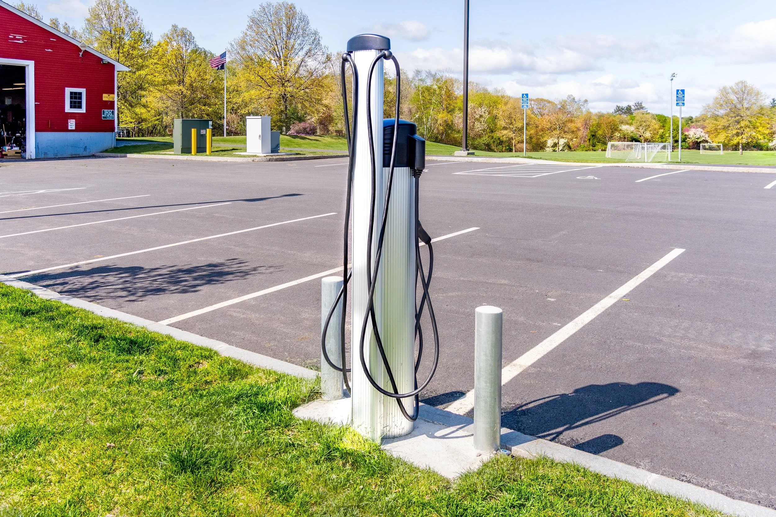 Electric vehicle charging station in an empty parking lot near a red building with a wide open door, surrounded by trees and a grassy area.