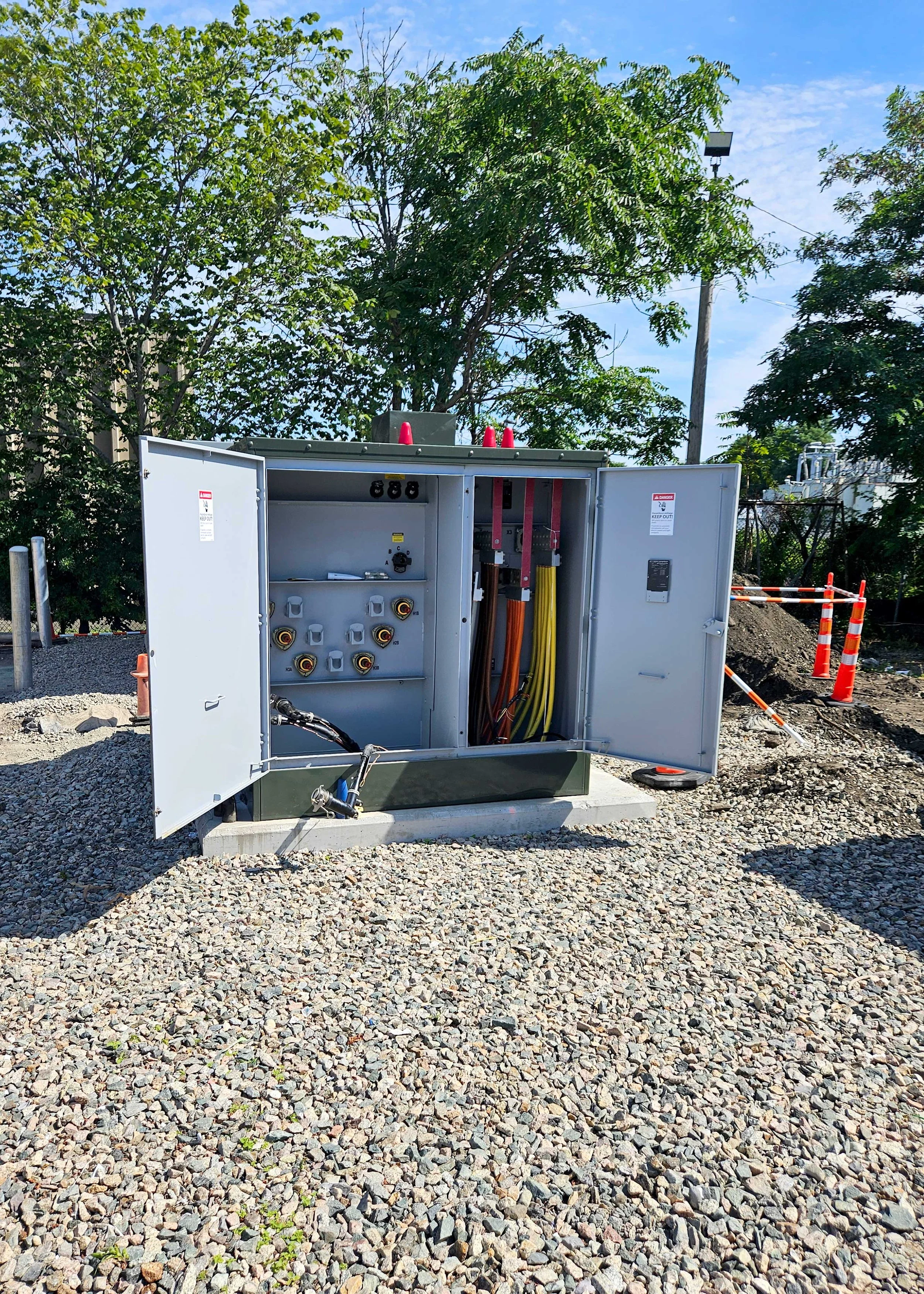 A large electrical or utility box with open doors revealing various wires, cables, and components inside, situated outdoors on a gravel surface with trees and a blue sky in the background.