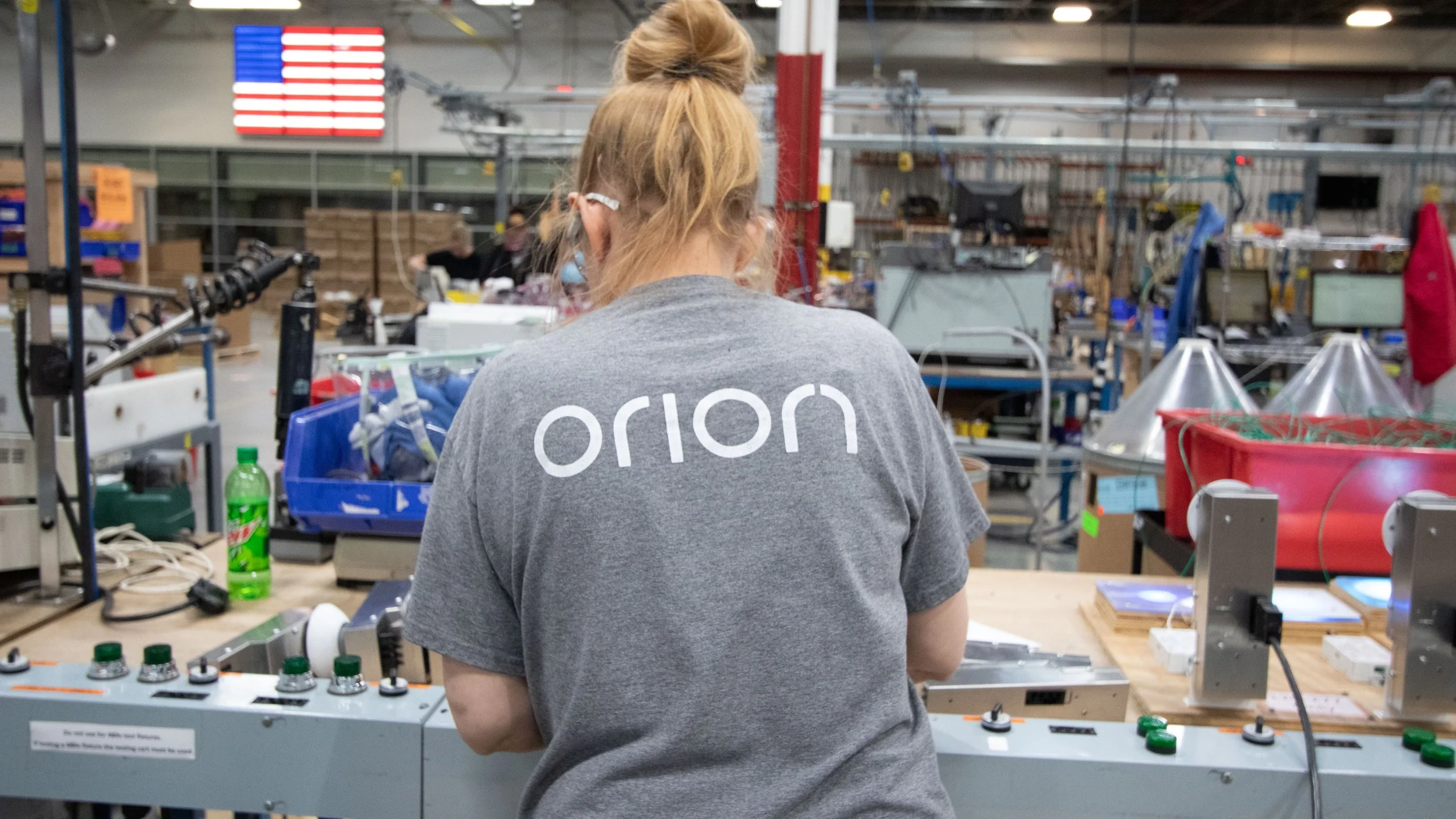 A woman stands at a workbench in an industrial or workshop setting, wearing a grey shirt with the word 'orion' on the back. Around her, various tools and electronic components are visible, with shelves and equipment in the background and a screen displaying an American flag.