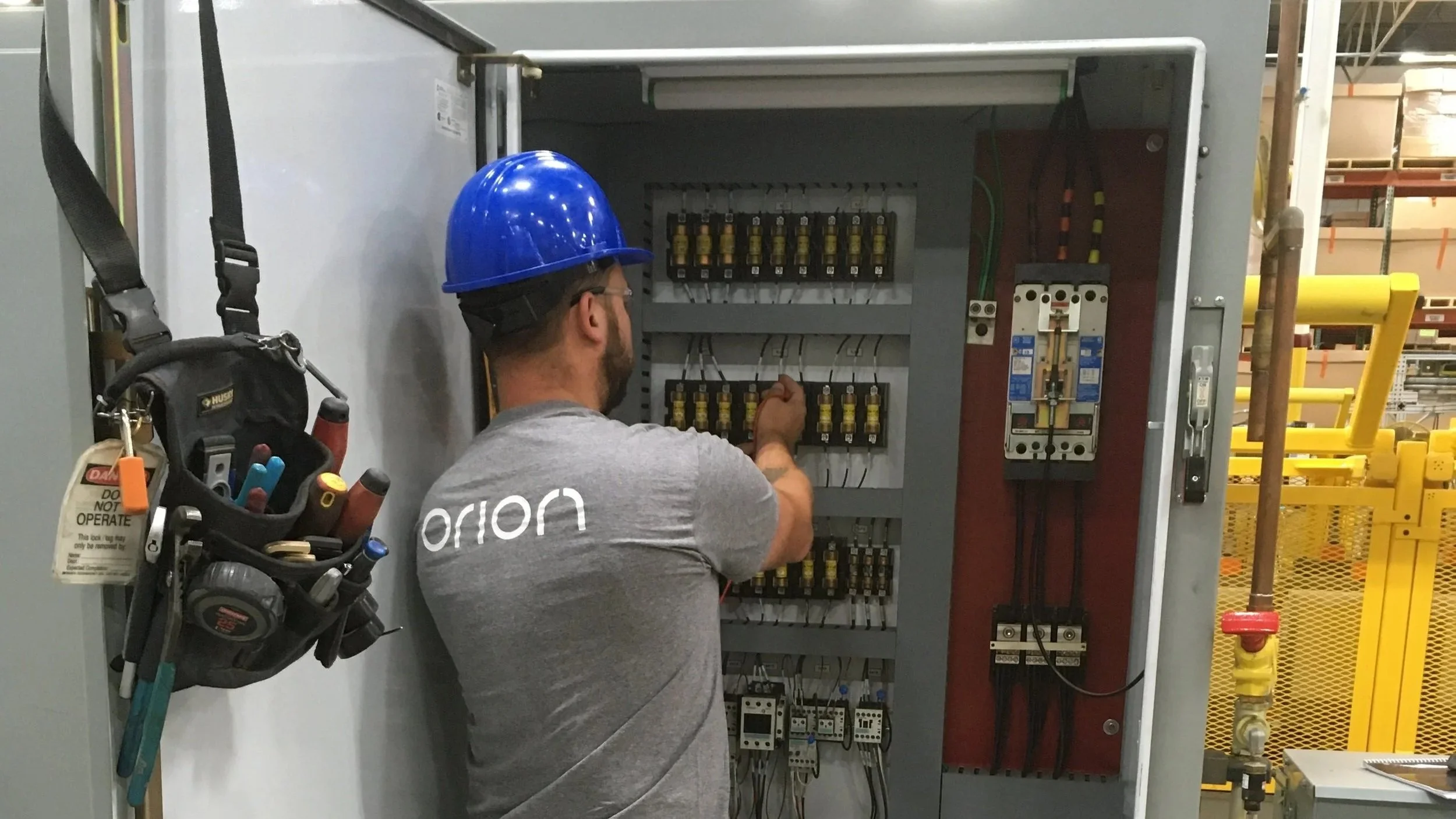 A worker wearing a blue safety helmet and gray shirt labeled 'orion' is working on an electrical panel with various wires, fuses, and circuit breakers inside an industrial setting.