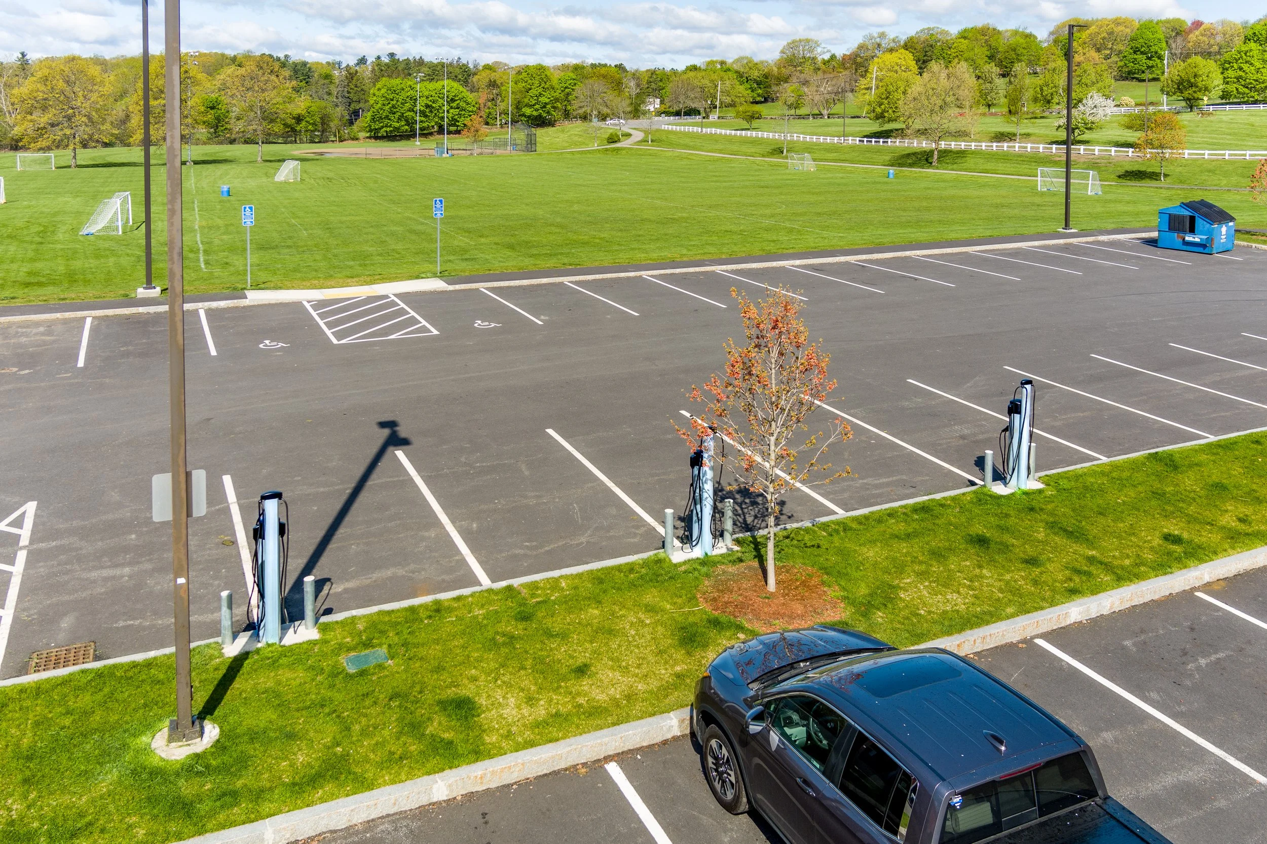 A parking lot with several electric vehicle charging stations, a black car parked, and a large green field with soccer goals and trees in the background.