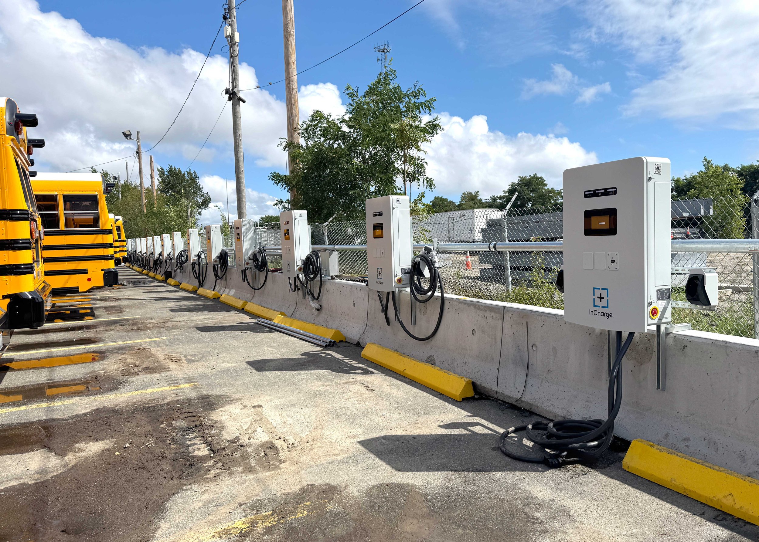 Electric vehicle charging station with multiple chargers installed outdoors next to a fence and a parked school bus, under a partly cloudy sky.