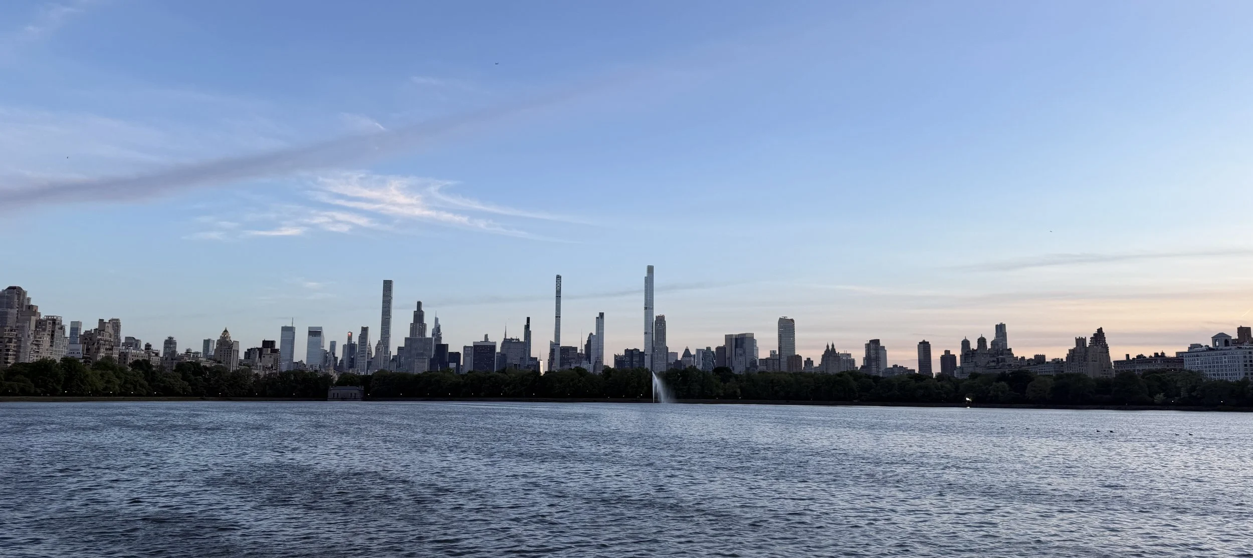 View of New York City skyline from Central Park Reservoir at sunset, with tall skyscrapers and a water fountain in the park.
