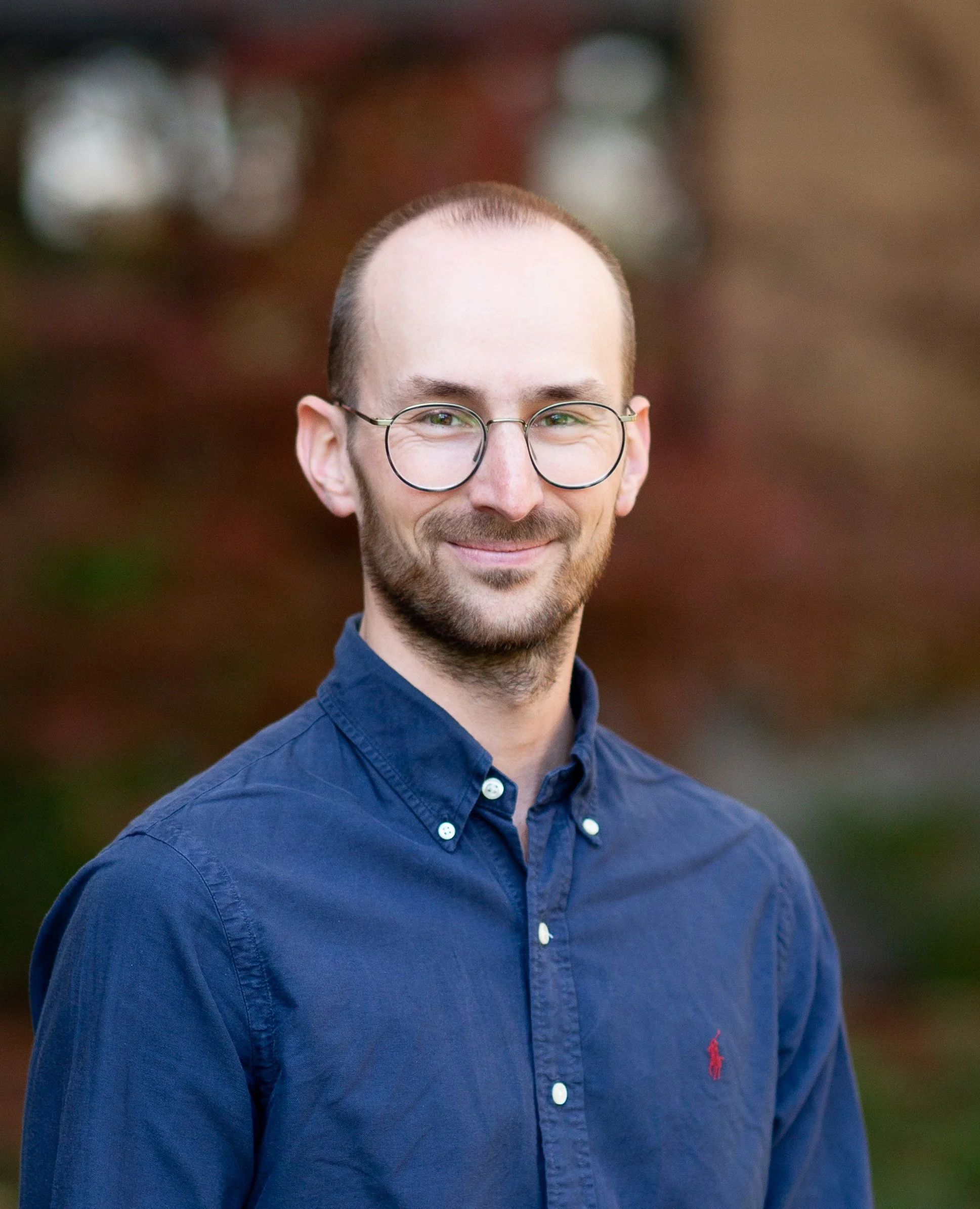 Close-up of a smiling man with glasses, wearing a blue button-up shirt, outdoors with blurred background.
