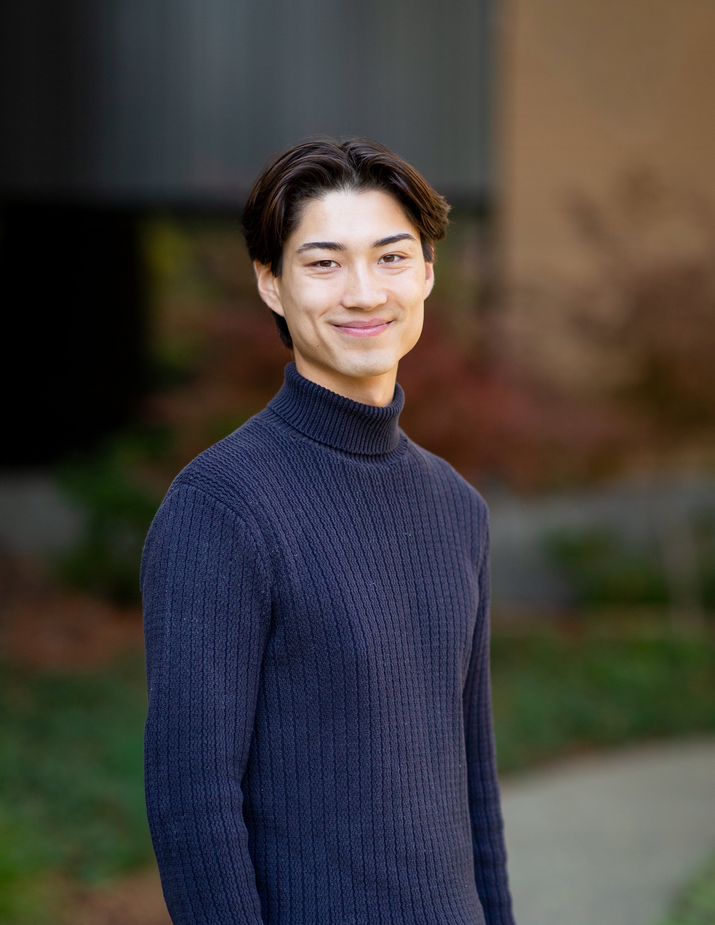 A young man with short dark hair wearing a navy blue turtleneck sweater standing outdoors with blurred trees and grass in the background.