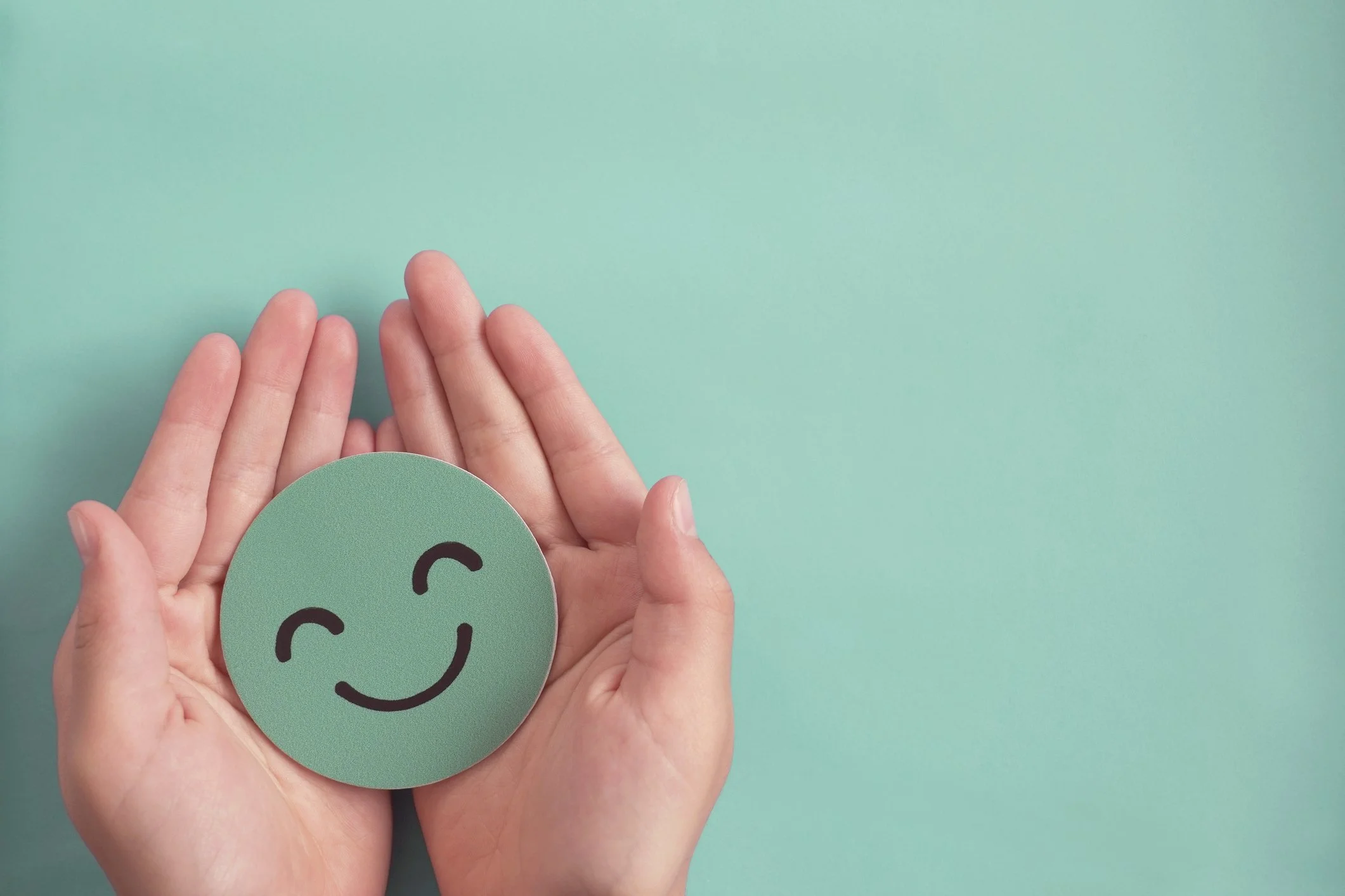 Close-up of human hands holding a green circular card with a smiling face and the letter C, against a light teal background.