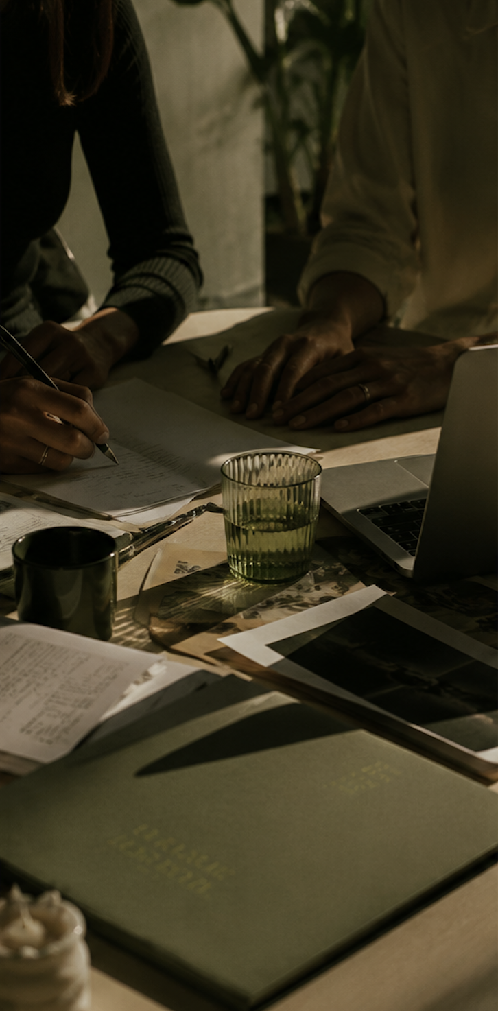 Two people working at a cluttered desk with papers, a laptop, a green glass of water, and a black mug, illuminated by warm lighting.