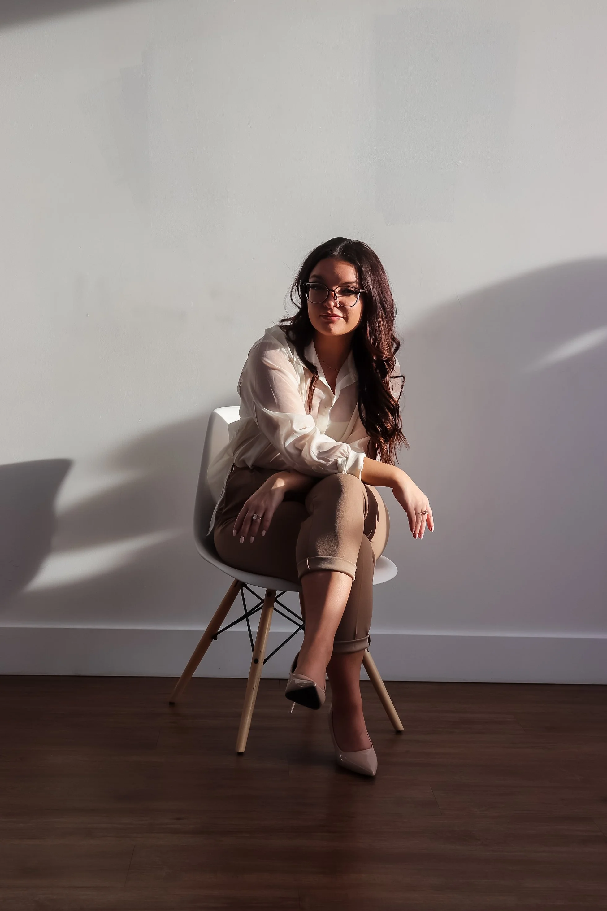 A woman with long dark hair, glasses, and wearing a beige blouse and pants, sitting on a white chair with wooden legs against a plain white wall. She has one leg crossed over the other and is wearing beige high heels.