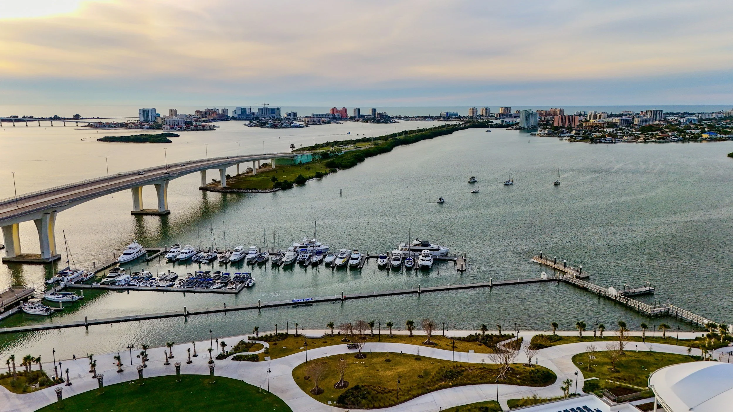 A view of a marina with several boats docked, a waterway, a bridge, and a city skyline in the background under a cloudy sky.