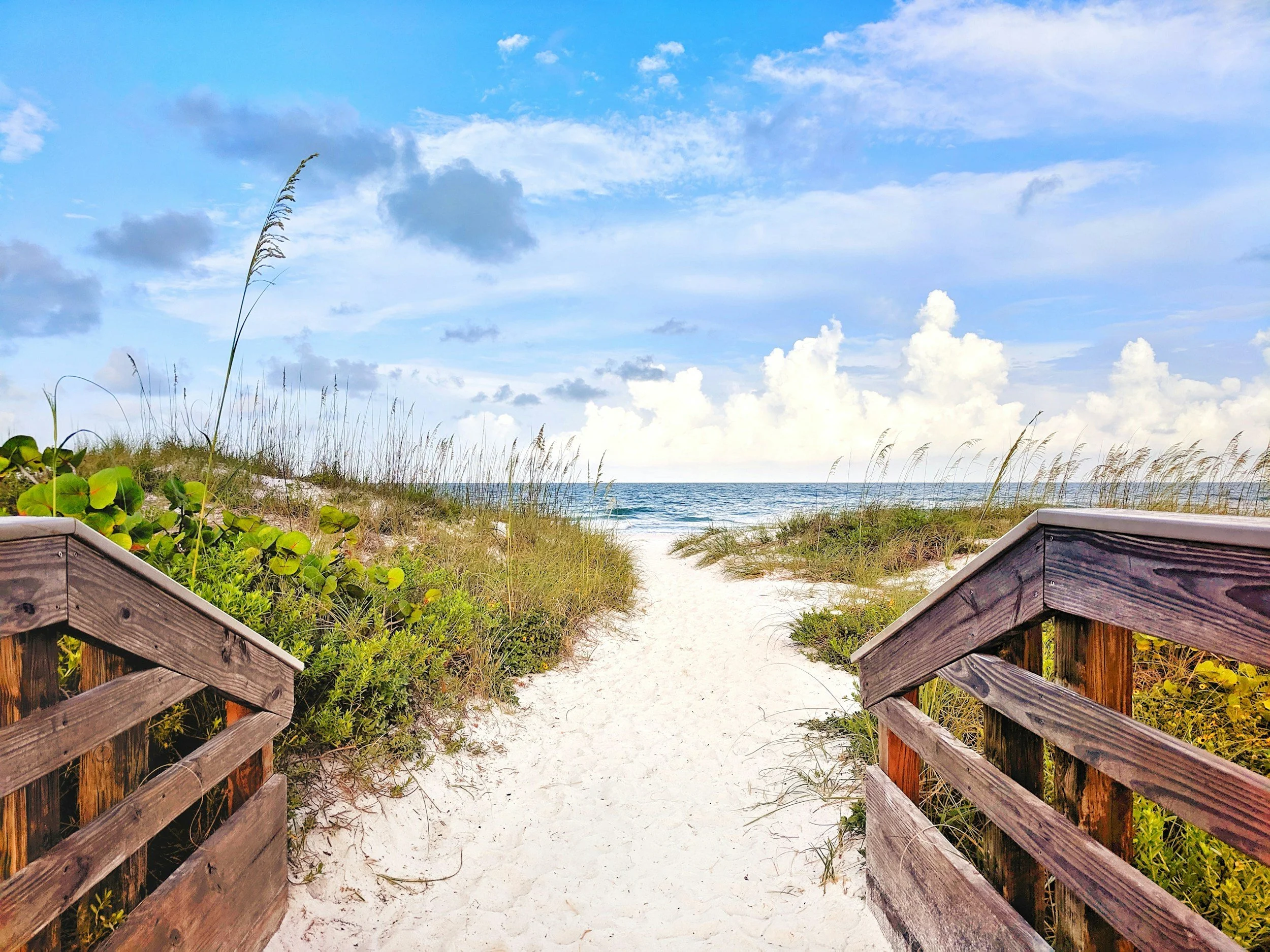 A sandy beach pathway bordered by wooden railings and lush green vegetation, leading towards the ocean with a partly cloudy sky overhead.