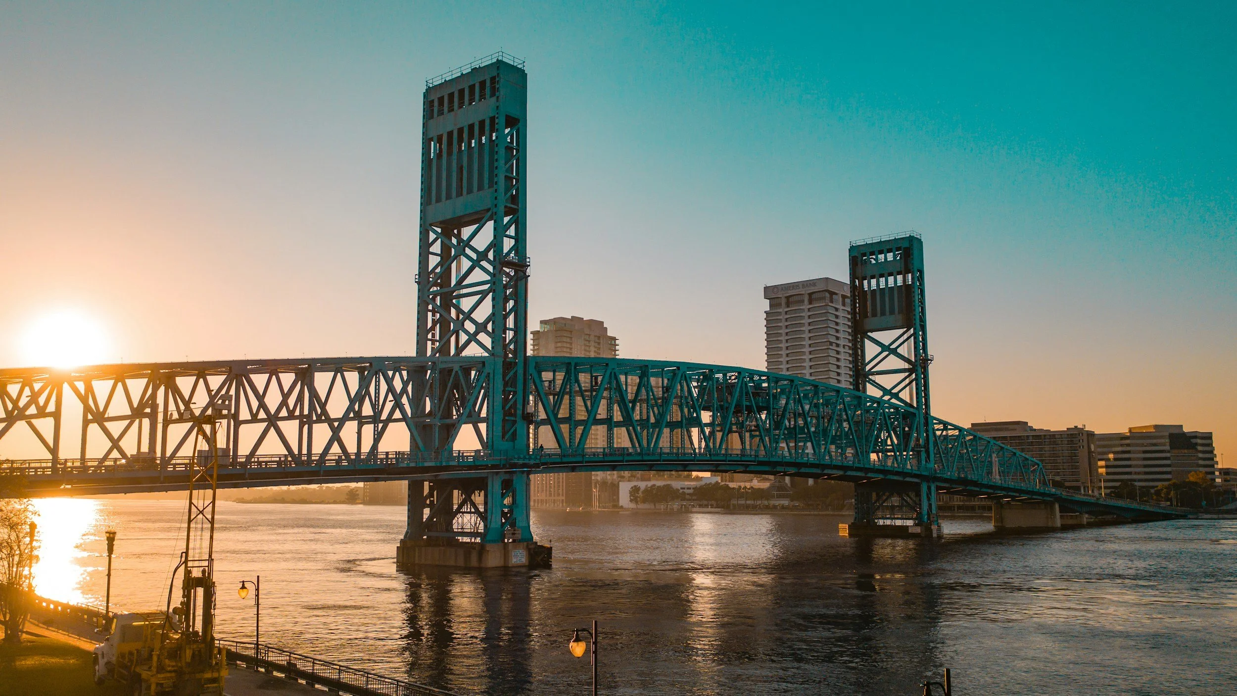 A blue steel bridge over a river at sunset with a city skyline in the background.